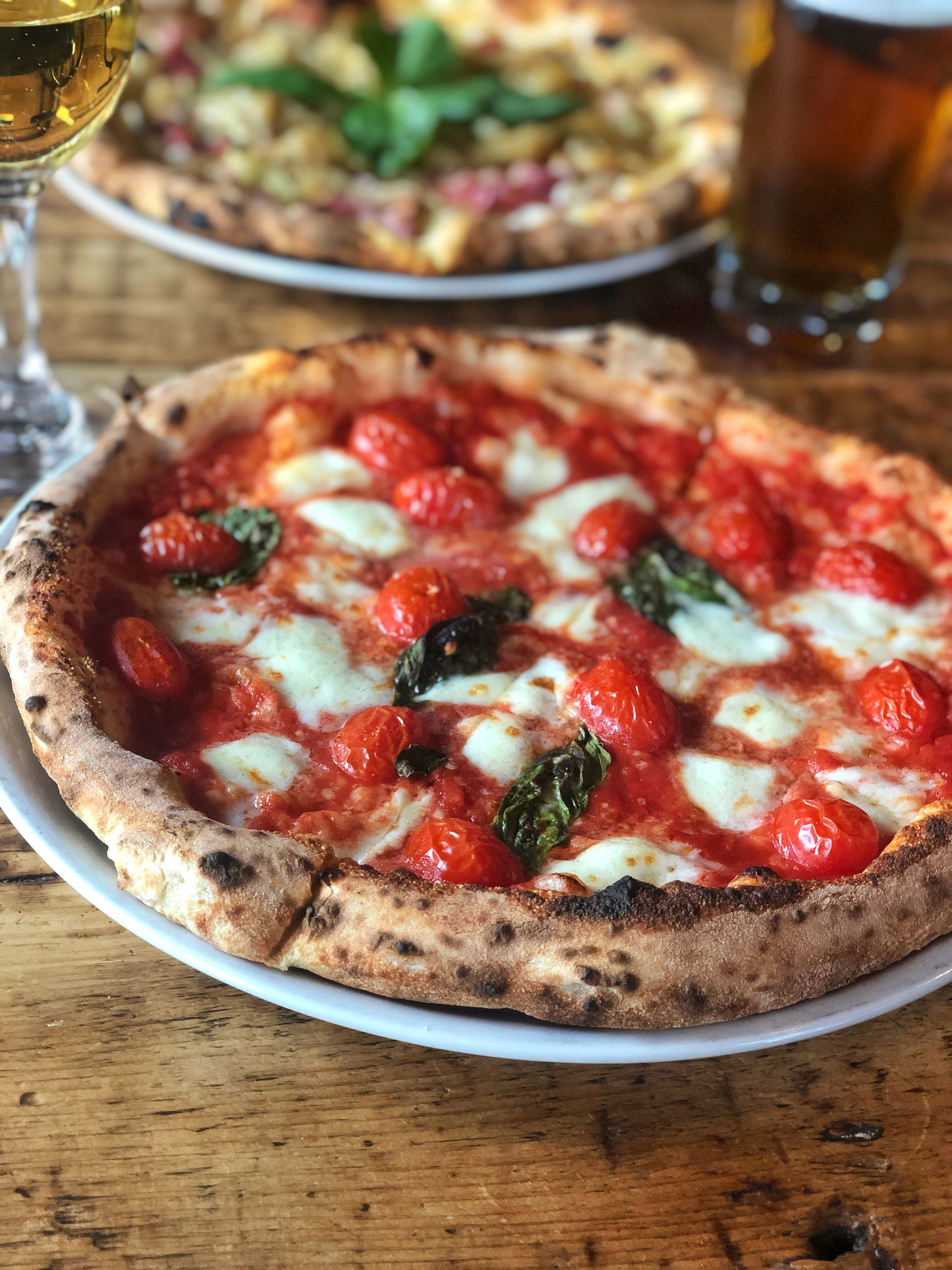 Close-up of a wood-fired pizza with cherry tomatoes, mozzarella cheese, and basil on a white plate. In the background, there is another pizza with green toppings and a glass of beer or cider on a wooden table.