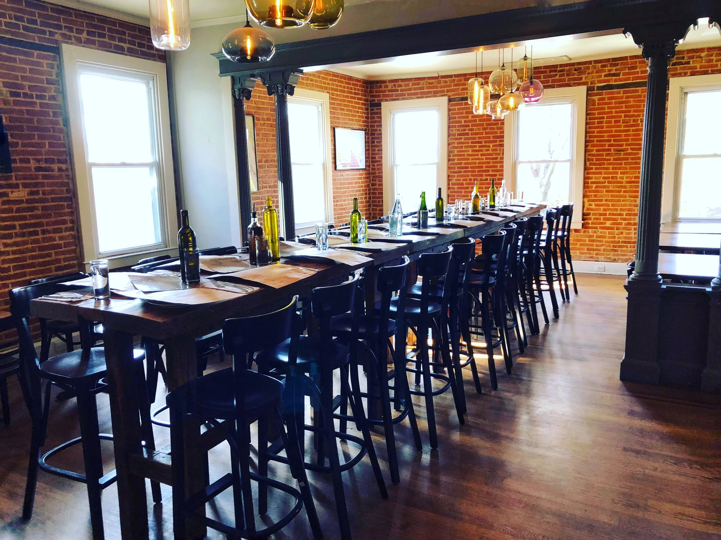 A long wooden dining table set with placemats and bottles of wine in a room with brick walls and large windows. There are black chairs around the table and modern pendant lights hanging from the ceiling.