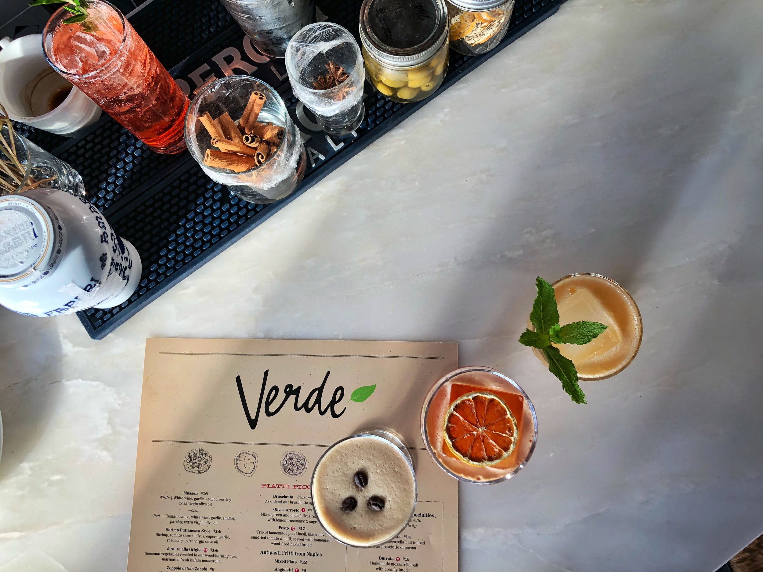 A menu card on a table with various drinks and jars of ingredients, including a drink garnished with mint, slices of dried citrus, and a sprig of mint, and several jars filled with spices and olives.