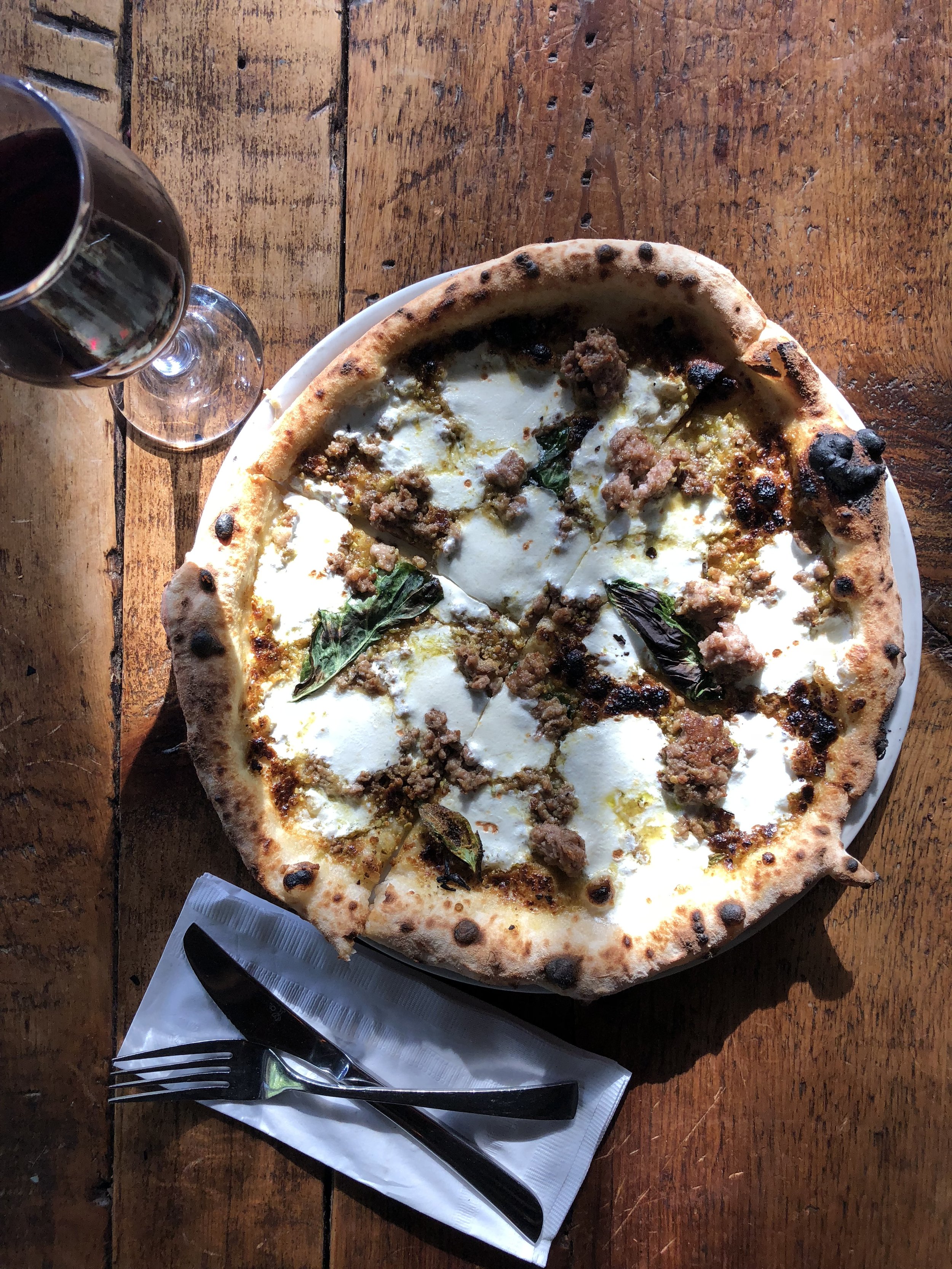 A pizza with sausage, basil leaves, and cheese on a wooden table, with a glass of dark soda and a fork and knife on a napkin.