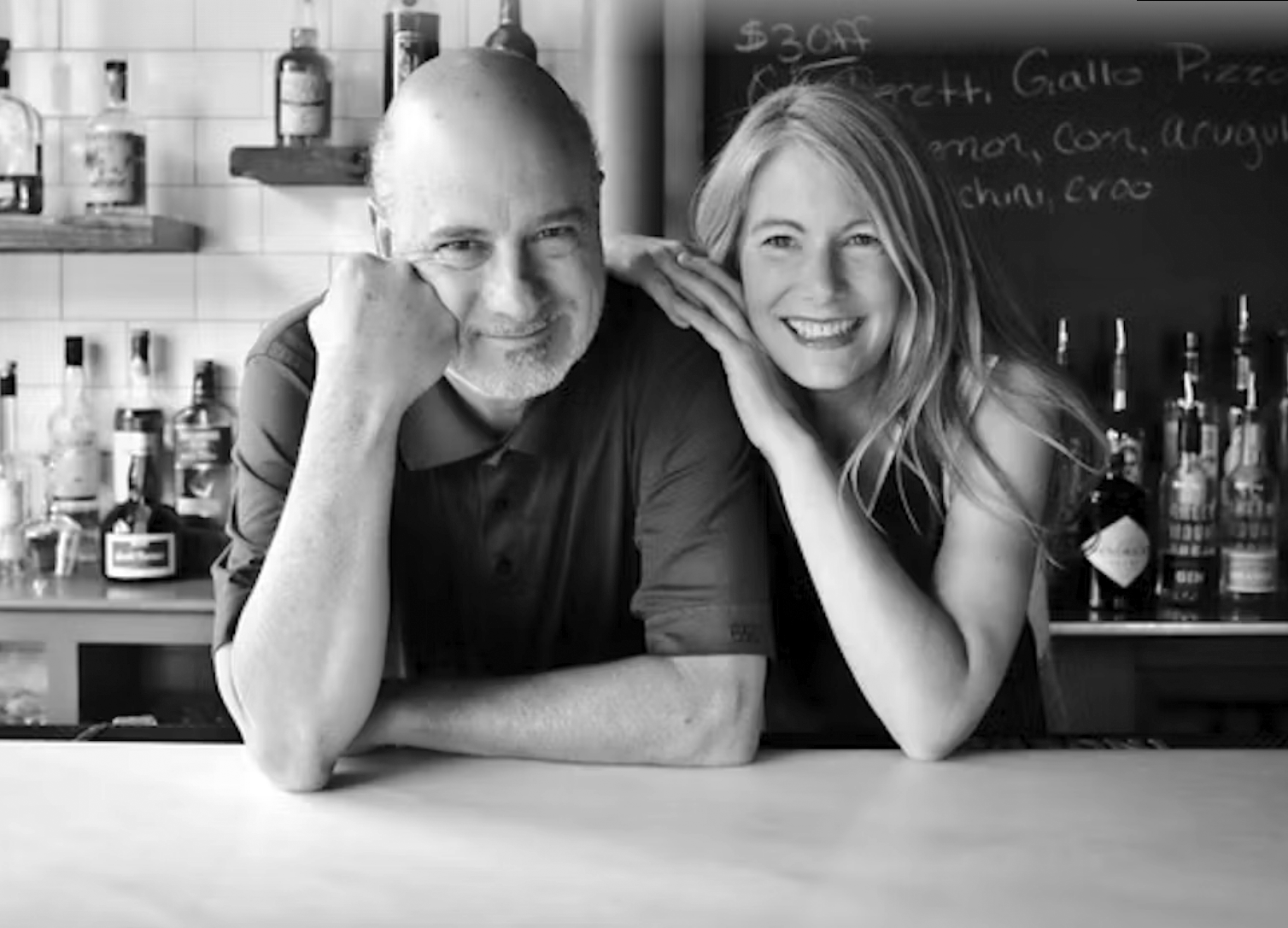 A black and white photo of a man and woman sitting at a bar, smiling, with bottles and a chalkboard menu in the background.