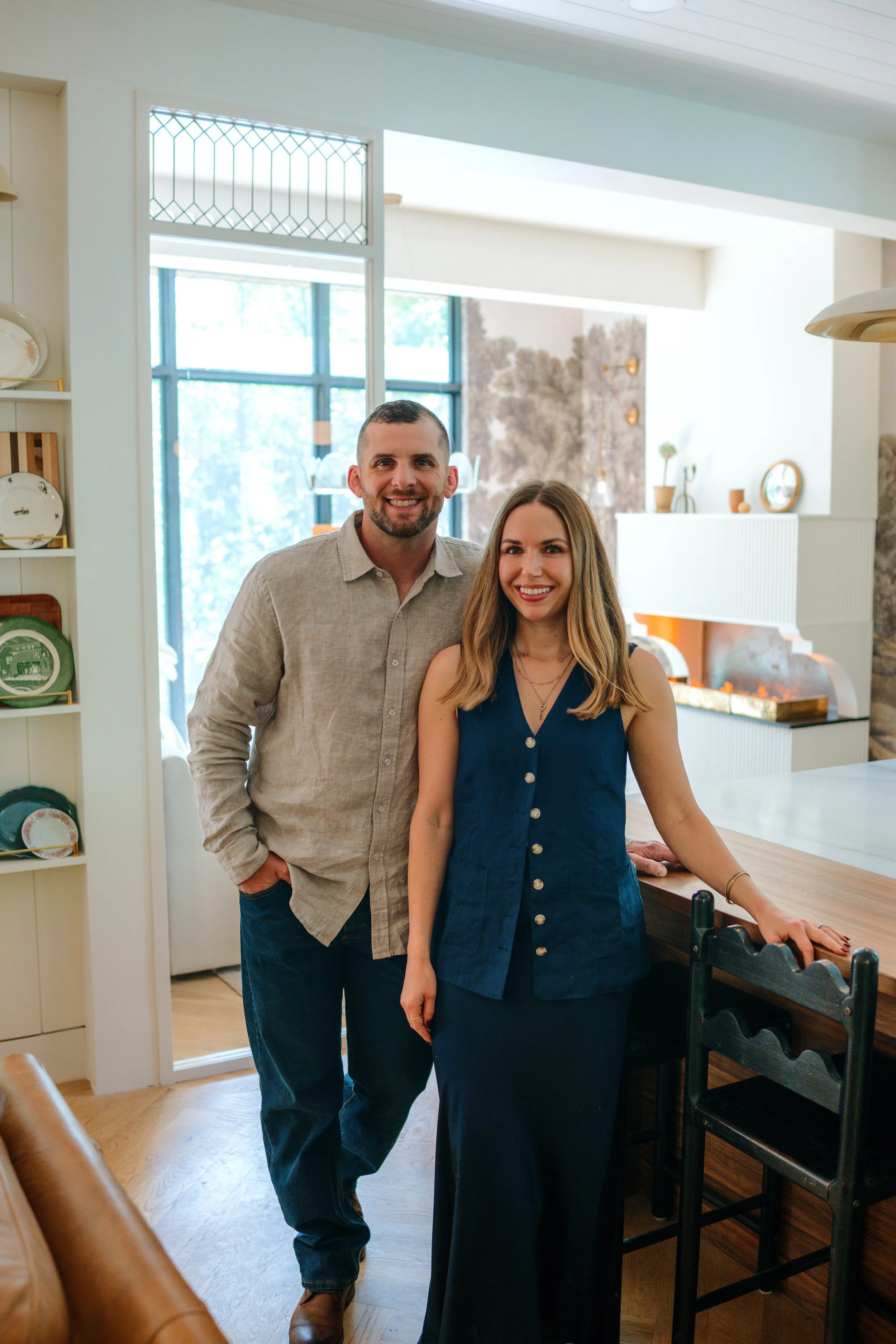 A man and a woman standing in a bright, cozy living room with large windows and modern decor, smiling at the camera.
