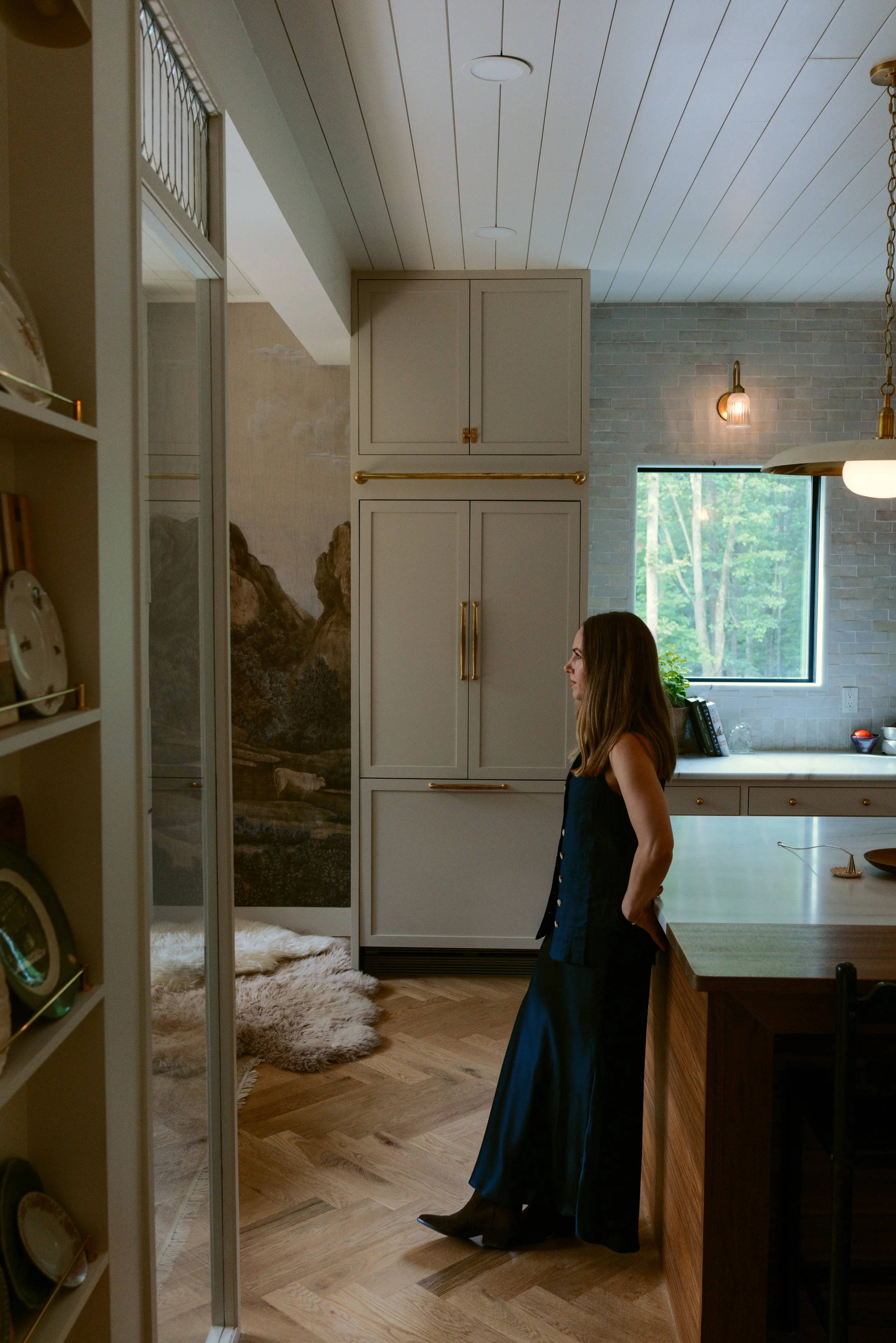 A woman with long brown hair in a black dress with buttons stands in a modern kitchen, near a wooden counter, with a window showing green trees outside. Inside, there is a large painting of mountains and trees on a wall, and warm lighting fixtures.