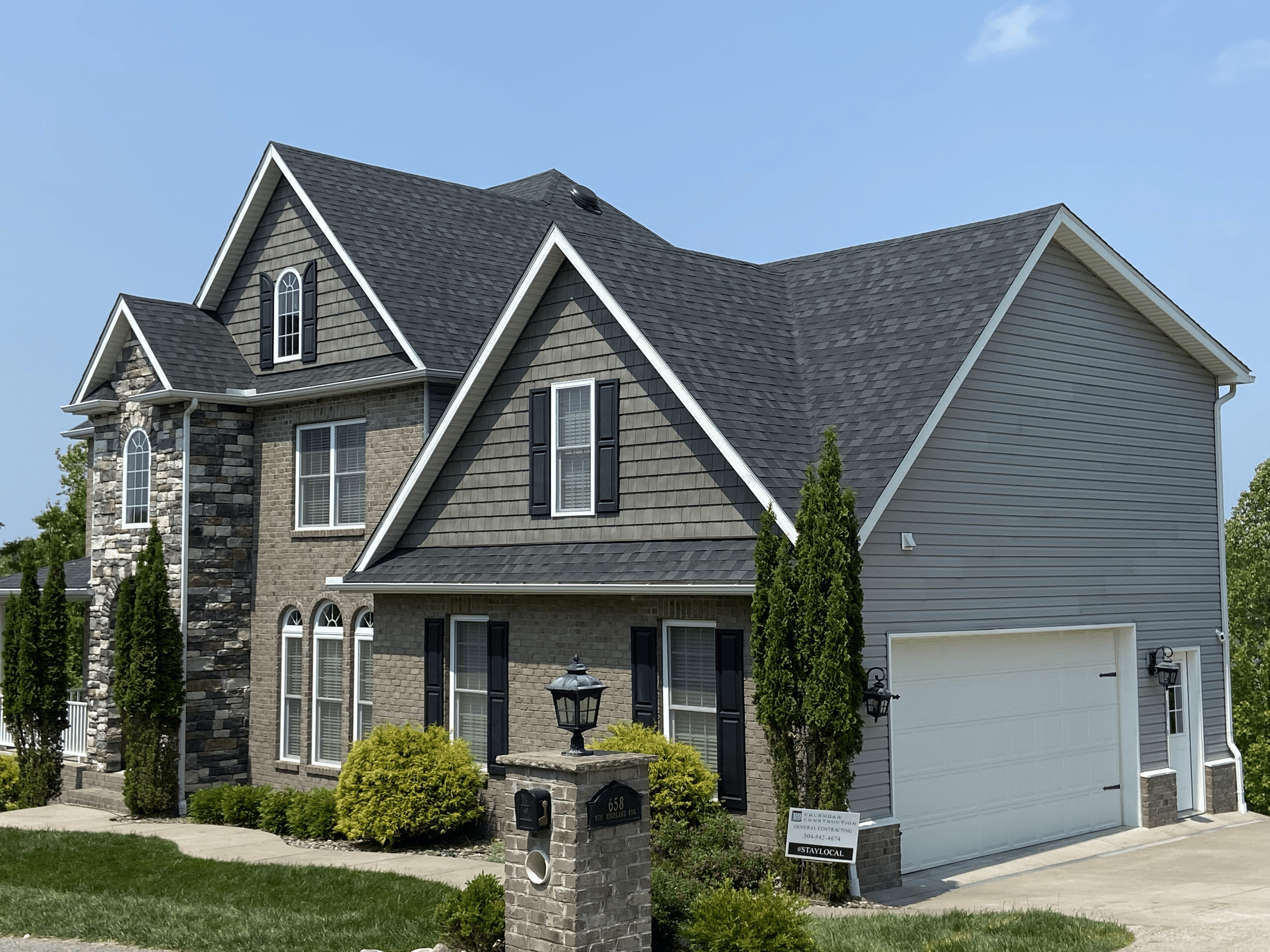 A large, multi-story house with gray roofing, brick and siding exterior, and several windows. The front yard has bushes and a mailbox on a brick pillar. Signs in the yard indicate construction and staying local.
