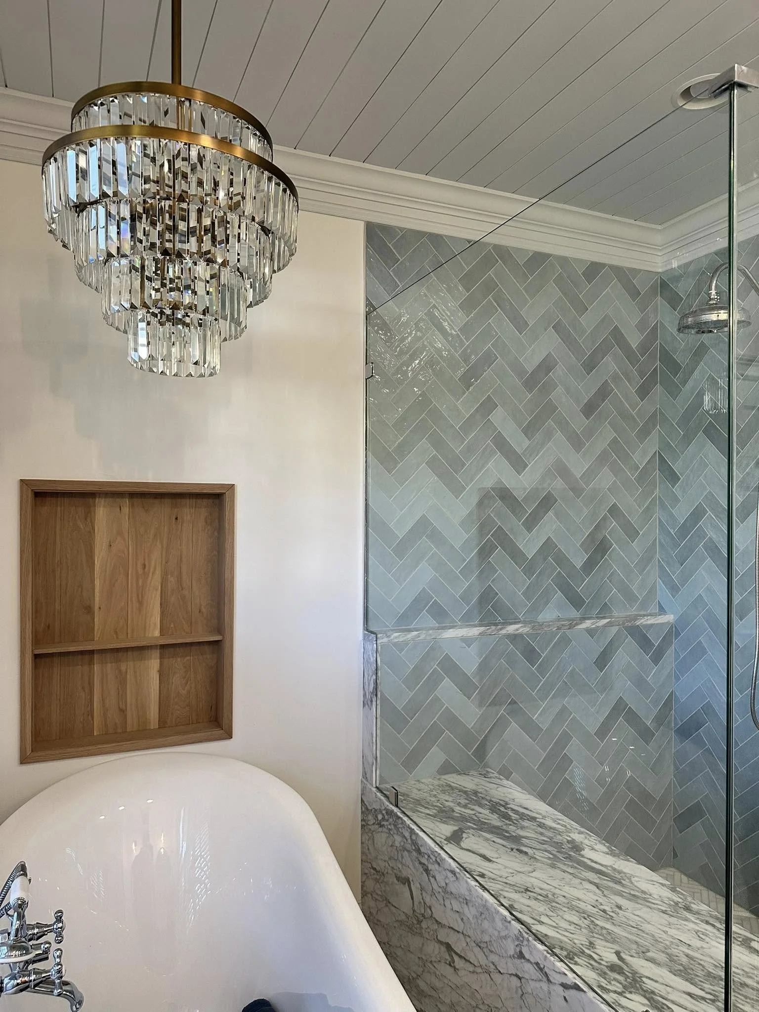 A bathroom with a large chandelier hanging from the ceiling, a built-in wooden shelving unit, a bathtub with chrome fixtures, and a walk-in shower with gray herringbone tile and marble base.