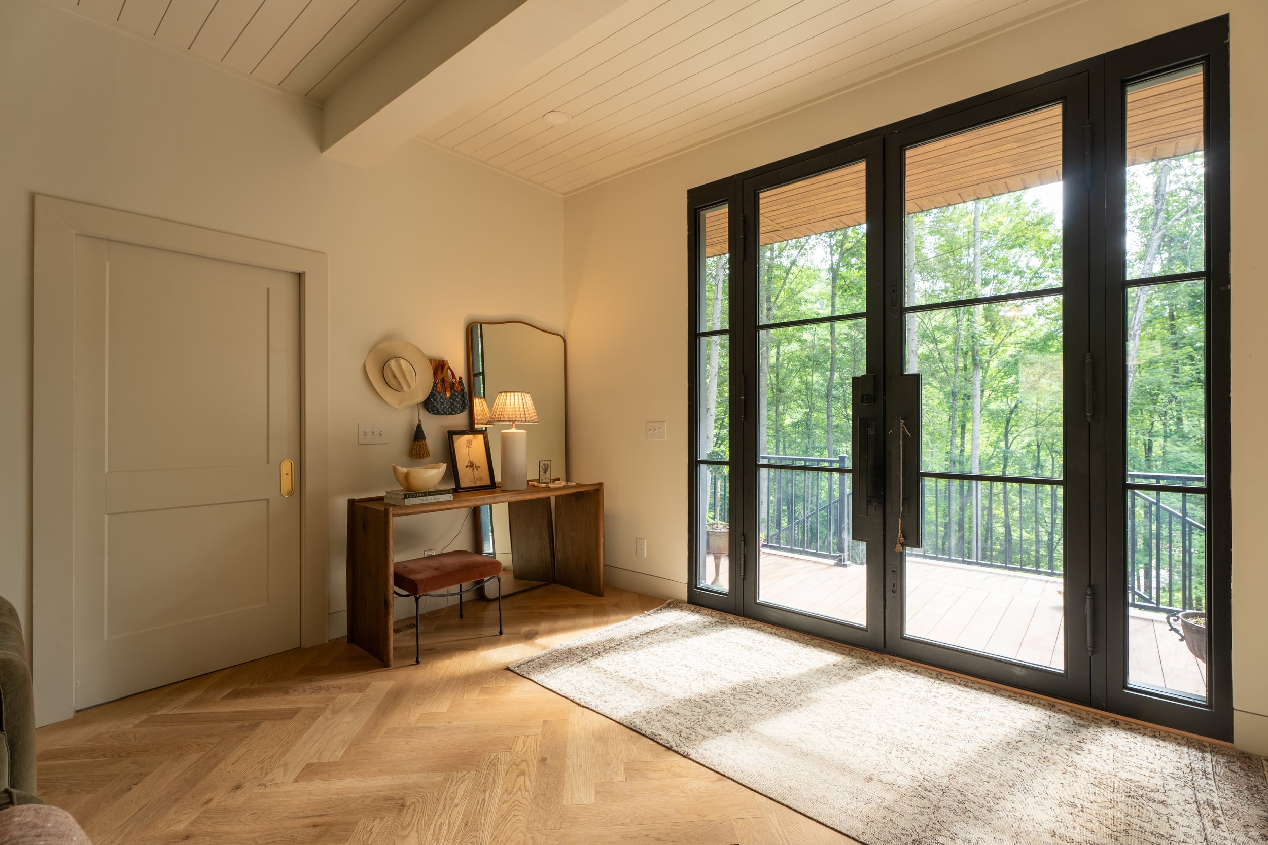 A cozy living space with large glass doors opening to a balcony and lush green trees outside. The room has a light-colored wooden floor, a small wooden desk with a mirror, table lamp, framed pictures, and a hat hanging on the wall. A beige rug is placed near the door, and an area of the ceiling is visible.
