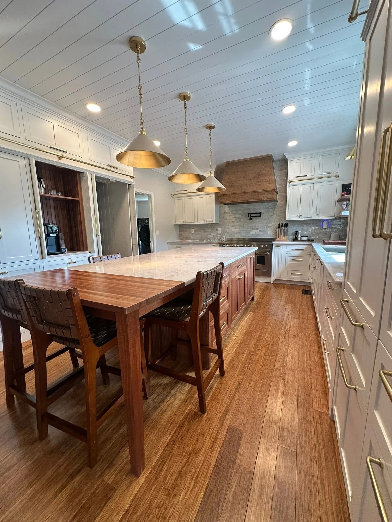 A modern kitchen with white cabinets, wooden accents, a large island, three pendant lights, wooden flooring, a brick stove hood, and stainless steel appliances.
