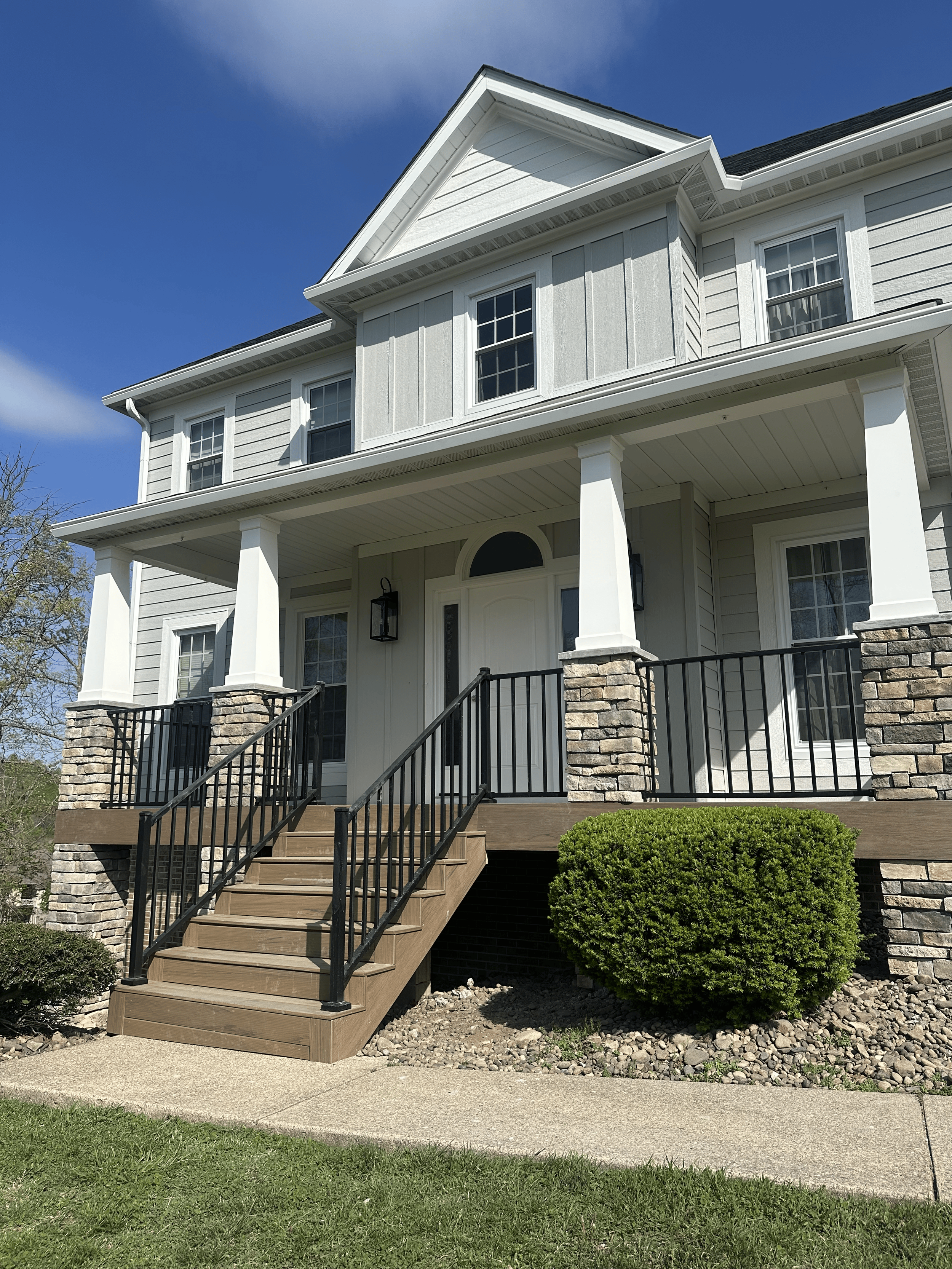 Front view of a modern two-story house with a porch, black railing, and stone pillars, surrounded by a well-maintained lawn and bushes, under a clear blue sky.