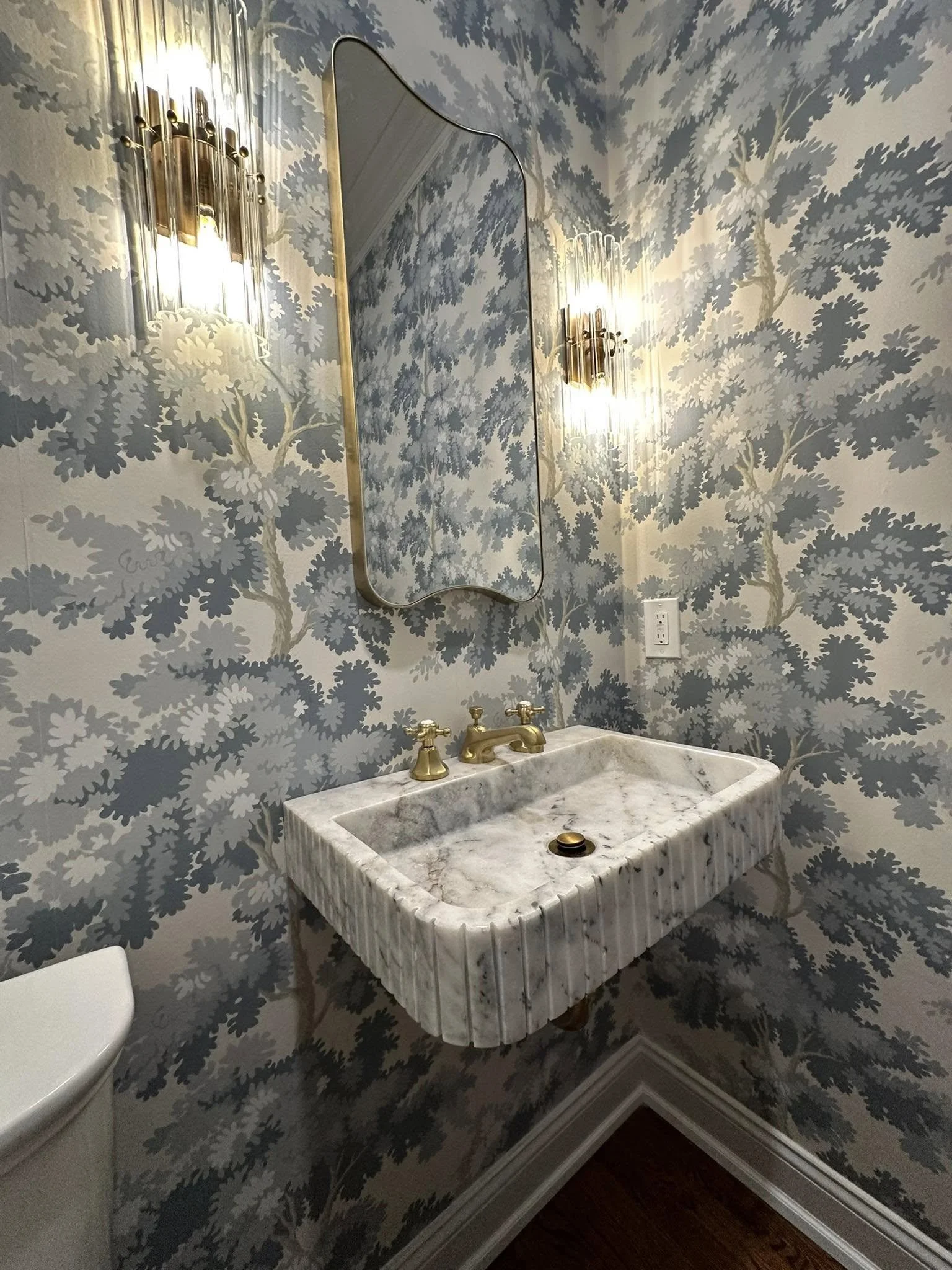 Bathroom with blue and white leaf-patterned wallpaper, marble sink with gold fixtures, oval mirror, wall-mounted light fixtures, and a visible electrical outlet.