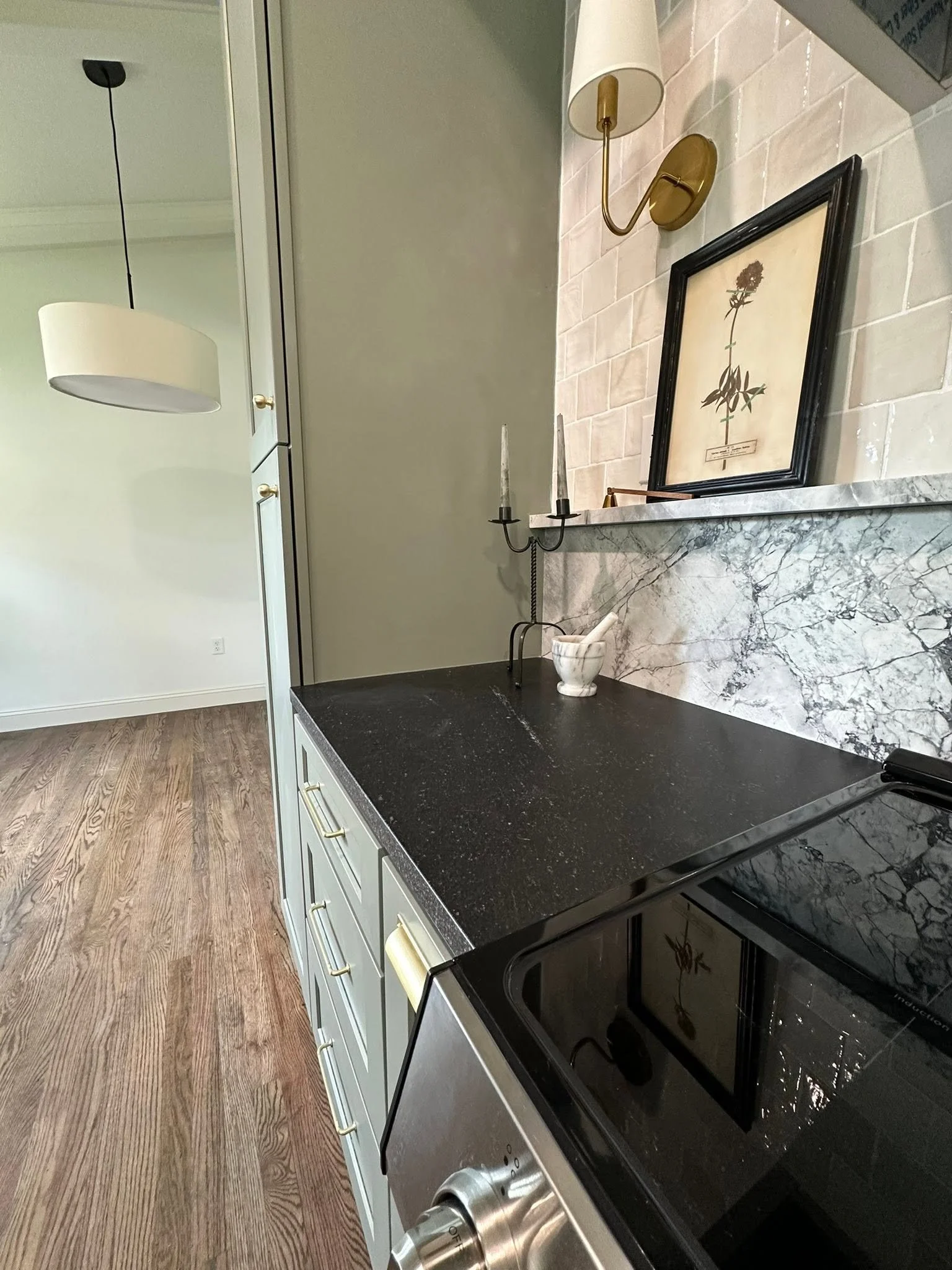 A kitchen counter with a black stovetop, white cabinets with gold handles, a marble backsplash, a small white bowl with a pestle, a brass wall-mounted lamp, and a framed botanical illustration of a plant.