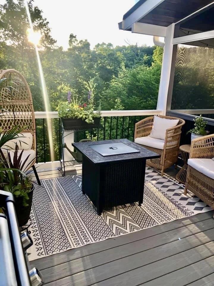 A cozy balcony with woven chairs and a small black table on a patterned rug, overlooking lush green trees with sunlight streaming through.