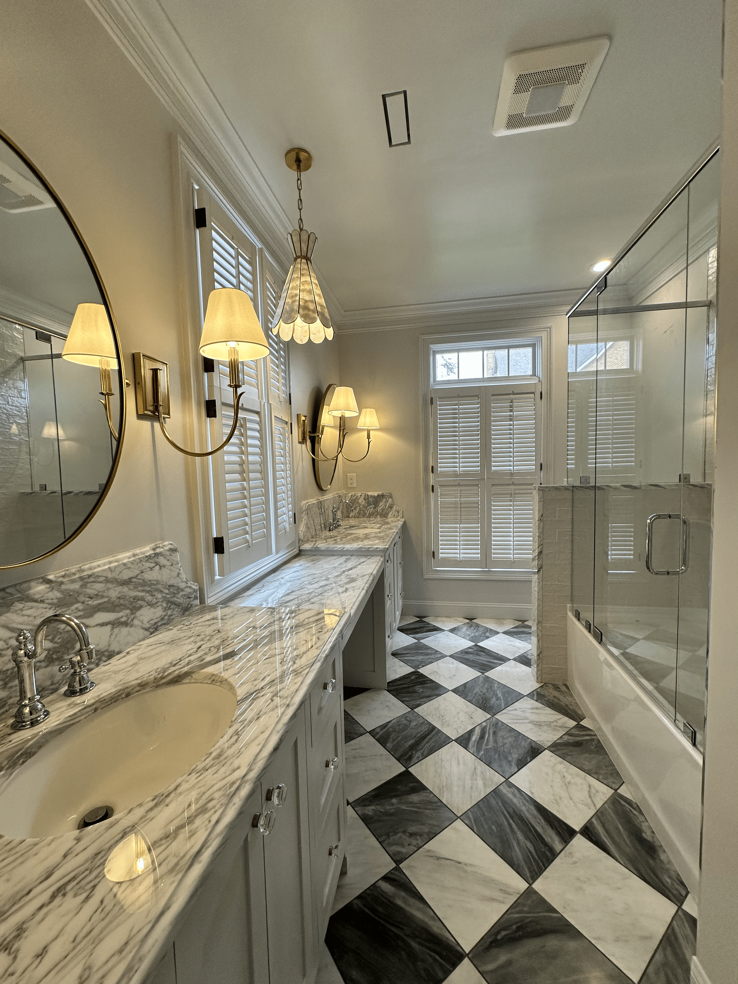 A luxurious bathroom with marble countertops and a patterned tile floor, featuring a round mirror, wall-mounted lamps, and a glass-enclosed shower.
