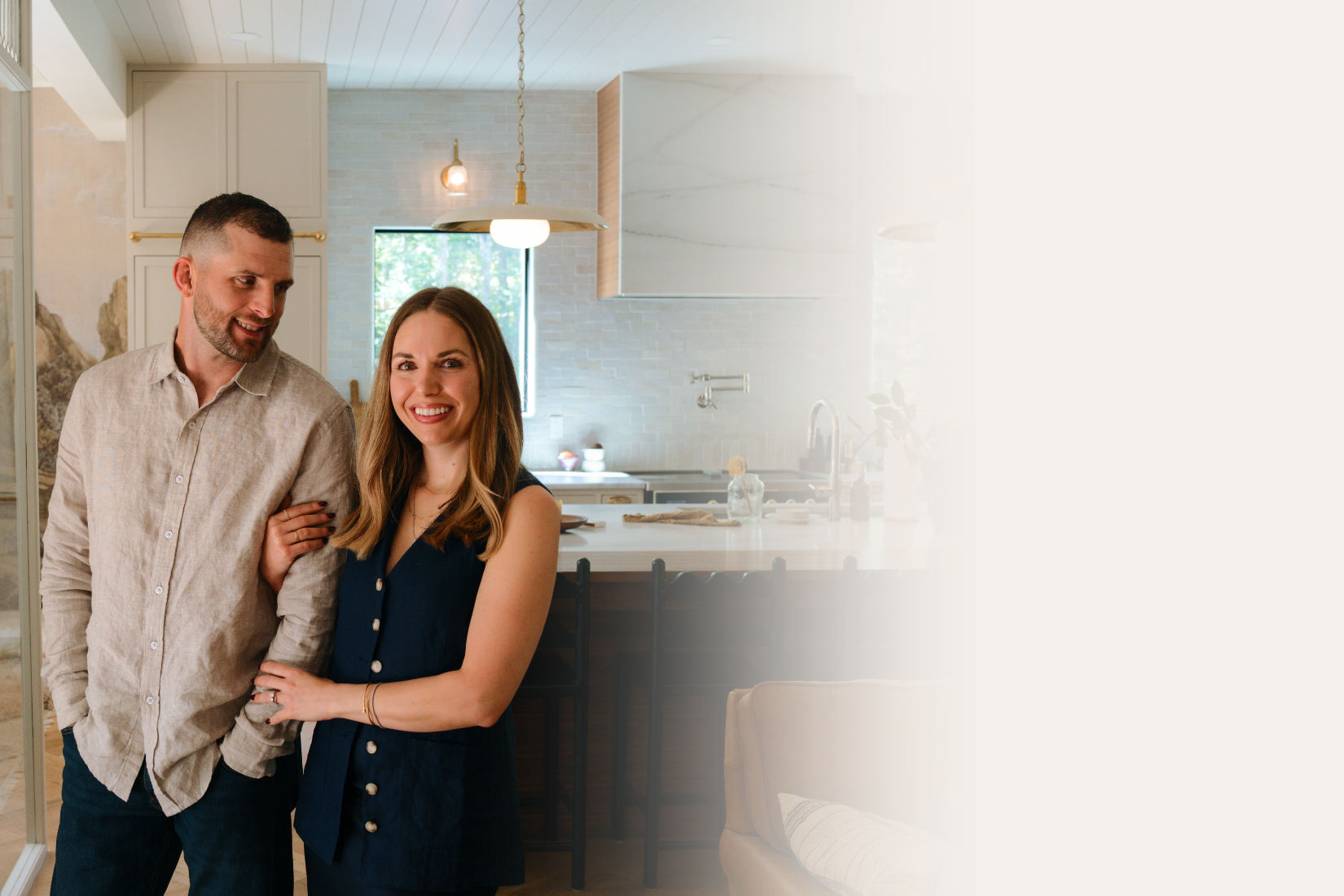 A smiling couple standing arm in arm in a modern kitchen with a white countertop, pendant light, and large window in the background.