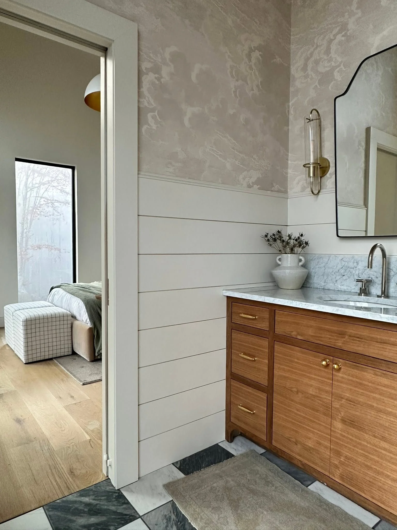 Interior view of a bathroom with a wooden vanity, marble countertop, a mirror, a brass wall-mounted light fixture, and a vase with dried plants. Part of a bedroom with a bed, pillow, and window is visible through an open doorway.