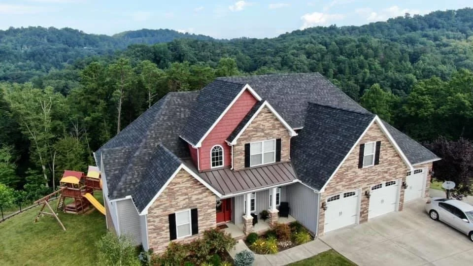 A large brick and siding house with a dark shingle roof, three-car garage, small front porch, and a backyard with playground equipment, set in a green, hilly landscape.