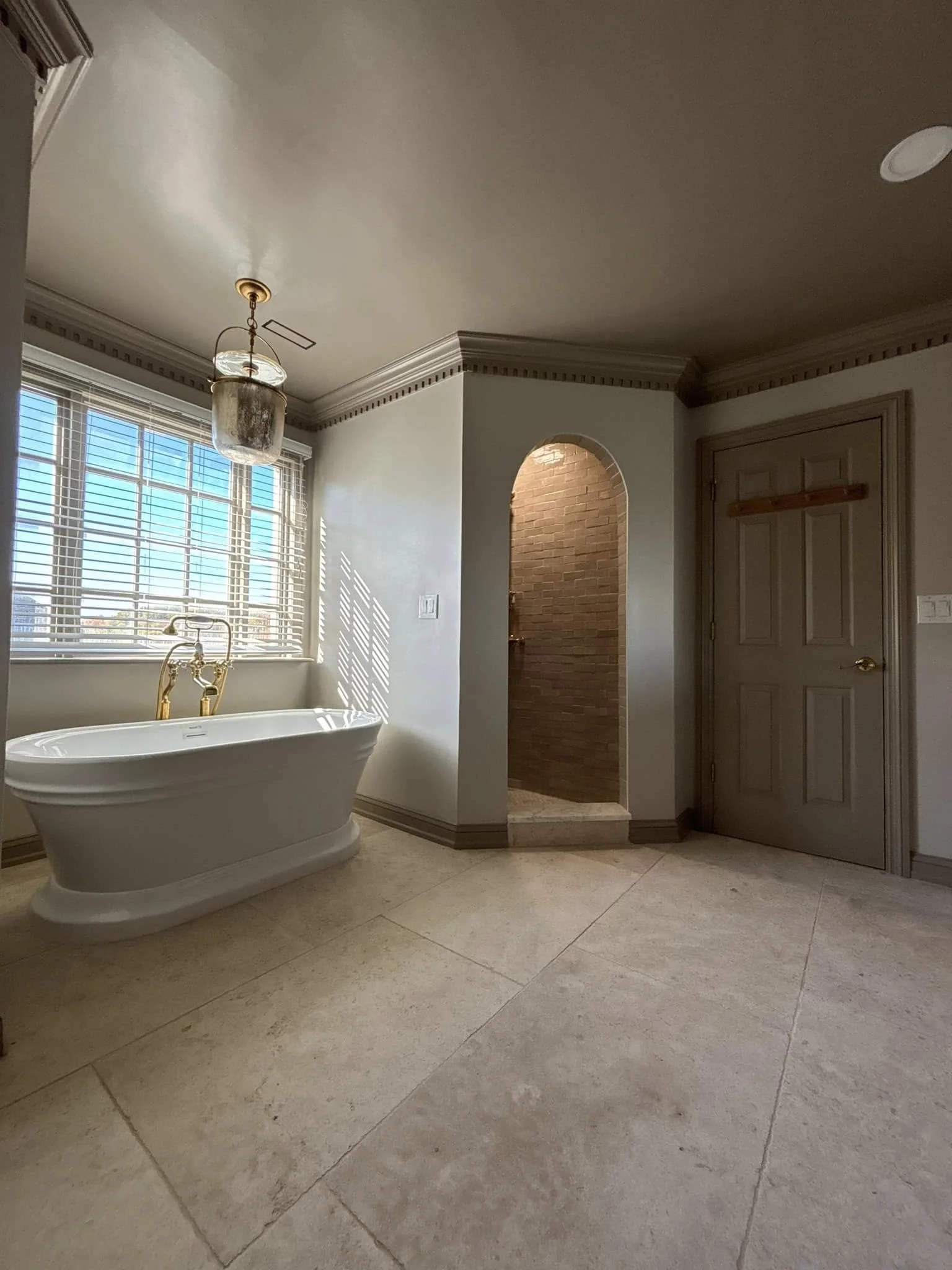 A bathroom with large beige tile flooring, a white bathtub with gold fixtures under a window with white blinds, and a small alcove with brick interior and arch, next to a closed grey door with gold handle.