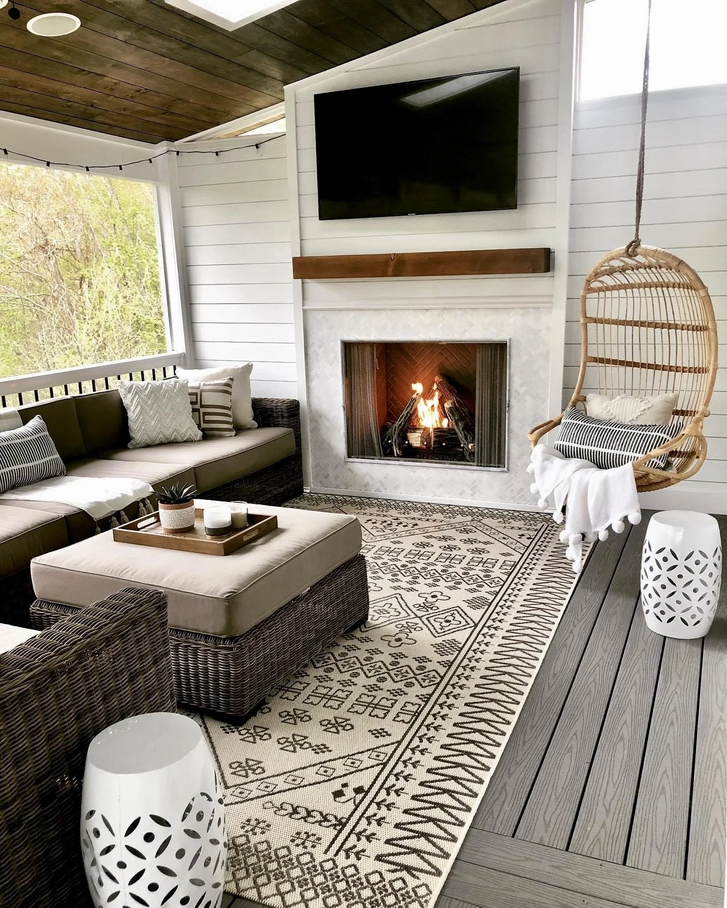 Living room with fireplace, wall-mounted TV, hanging wicker chair with pillow, green sofa with patterned pillows, patterned rug, black wicker furniture, white decorative lamps, wooden deck floor, and large window with greenery outside.