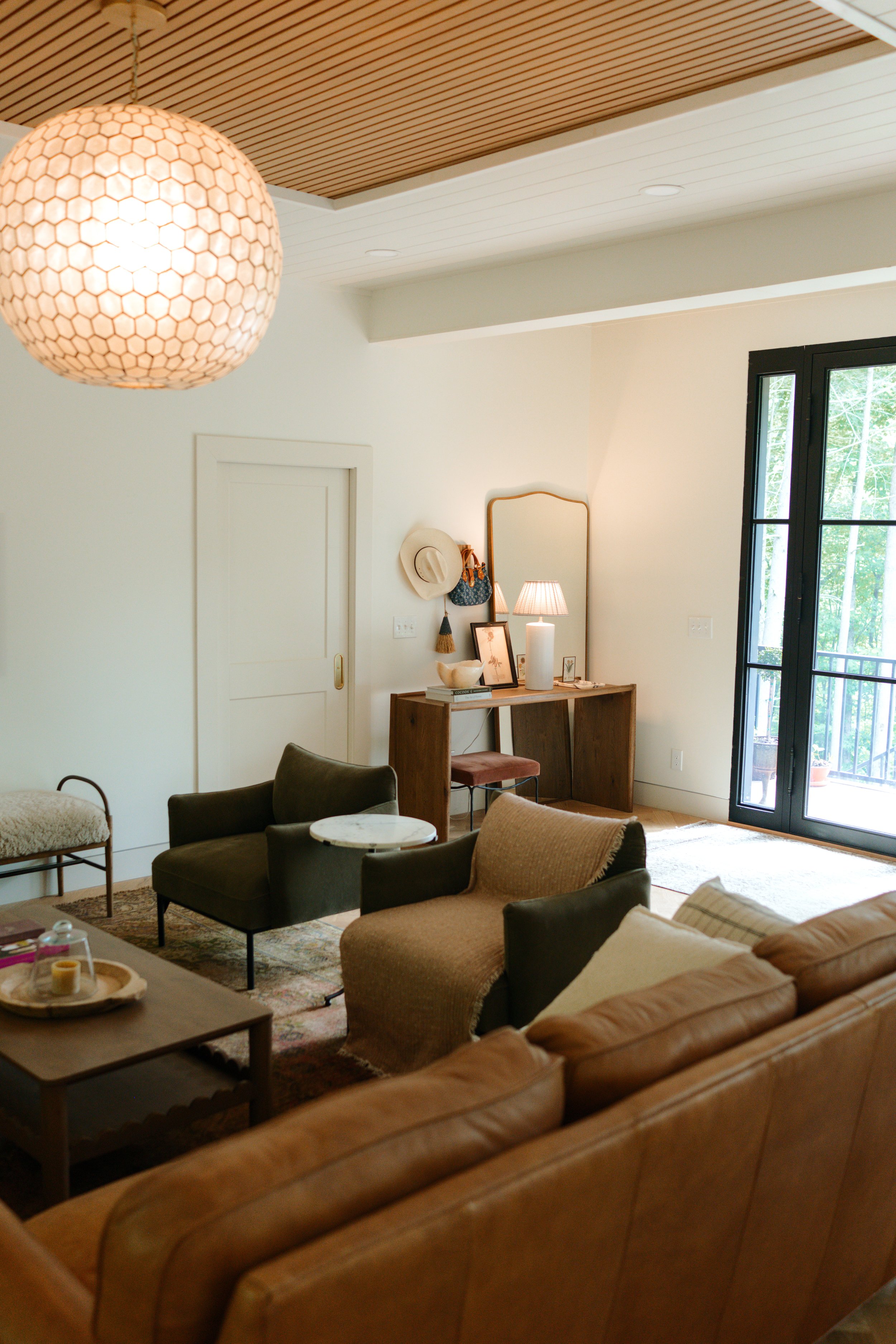 Living room with beige and brown sofas, a wooden console table with decorative items, a window with greenery outside, a hanging light fixture with a honeycomb pattern, and a wooden ceiling.