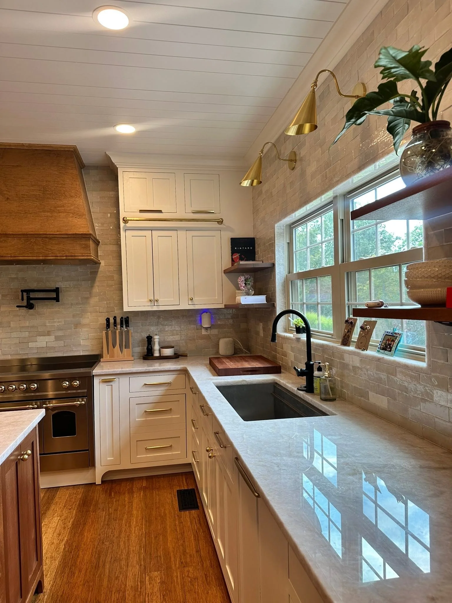 A kitchen with white and wood cabinets, a brick backsplash, a large window with a view of greenery, and a granite countertop. There are gold-colored wall sconces, a vase with greenery, and various kitchen items on the counter and shelves.