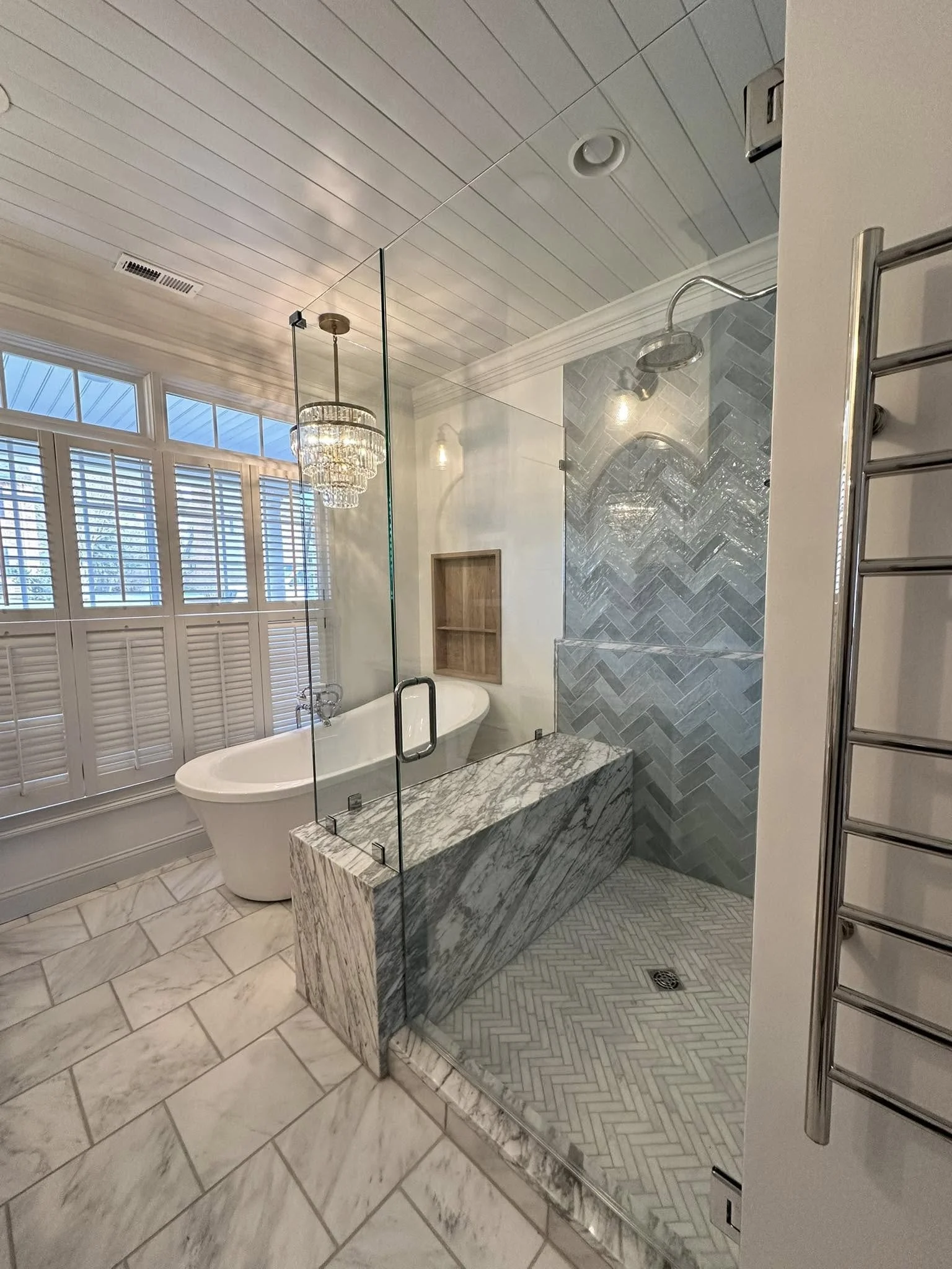 A modern bathroom with a walk-in shower with herringbone gray tile, a clawfoot bathtub, a chandelier, and white plantation shutters on the windows.