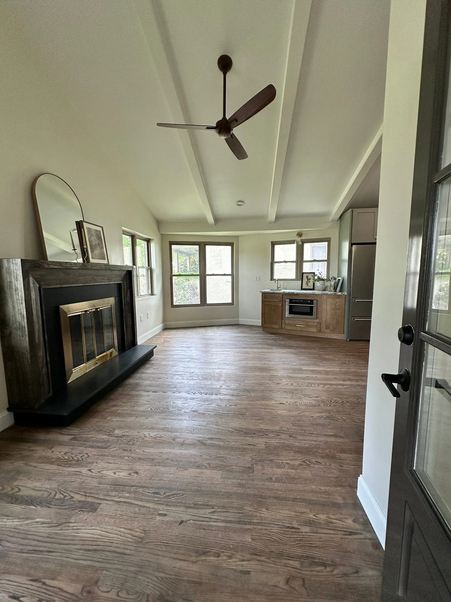 Spacious living room with wood flooring, fireplace, and multiple windows, kitchen with wooden cabinets, stainless steel refrigerator, and microwave visible in the background.