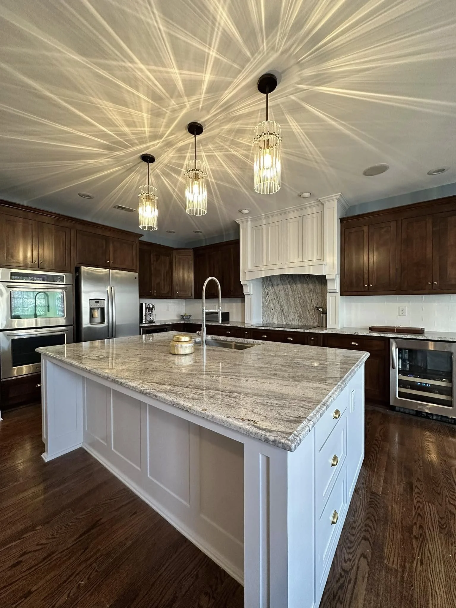 Modern kitchen with white kitchen island, dark wood cabinetry, stainless steel appliances, granite countertops, and pendant lighting casting starburst patterns on the ceiling.
