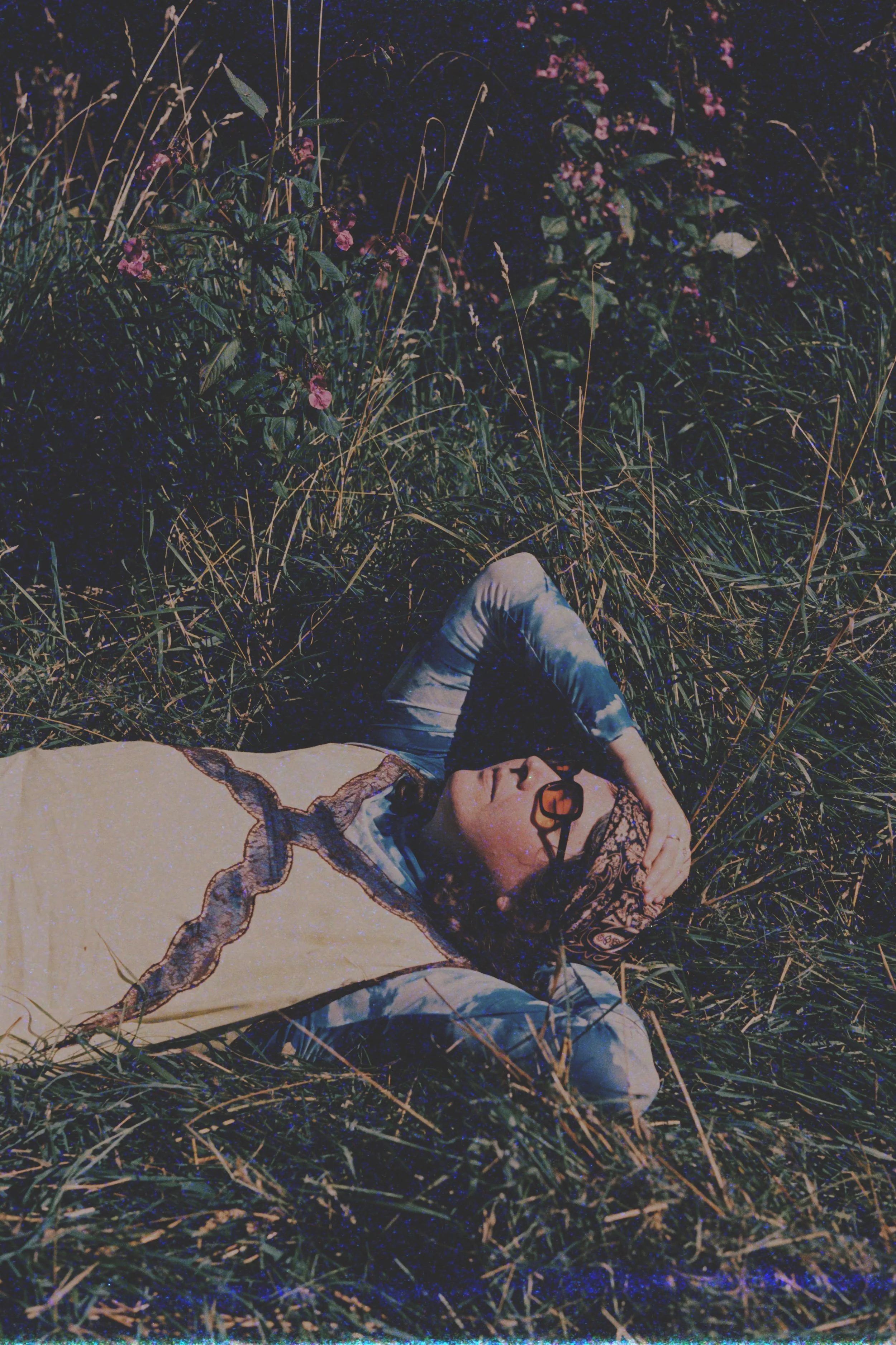 A woman lying on the grass at night, wearing glasses, a headscarf, and a beige top with lace details, surrounded by grass and small pink flowers.