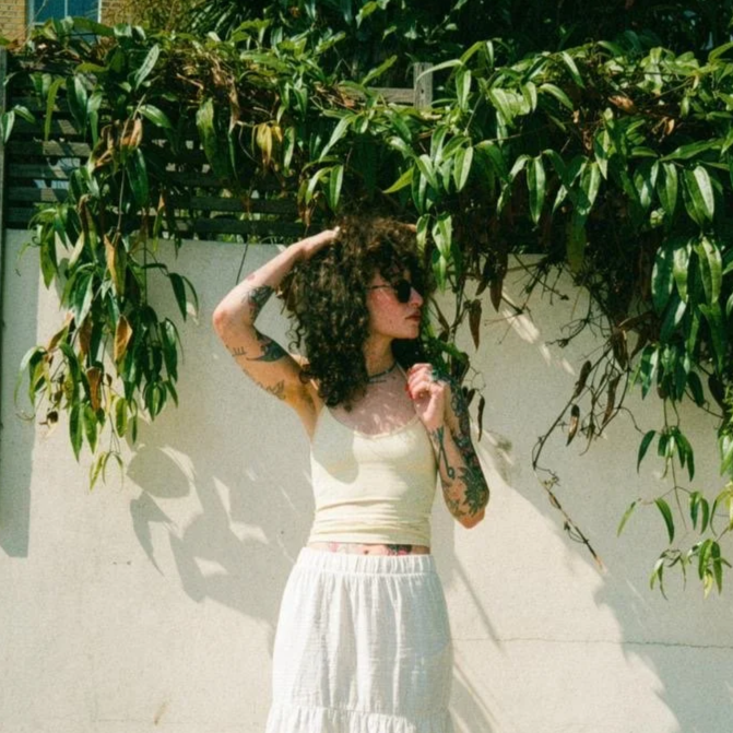 A woman with curly hair and tattoos wearing sunglasses, a beige tank top, and a white skirt stands in front of a white wall and green leafy plants, touching her hair with one hand and holding a flower with the other.