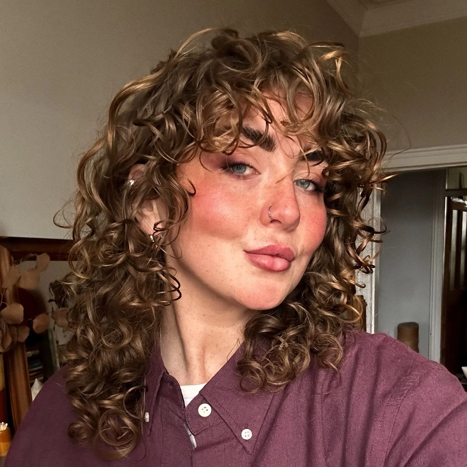 A woman with curly light brown hair and blue eyes wearing a maroon shirt, taking a selfie indoors.
