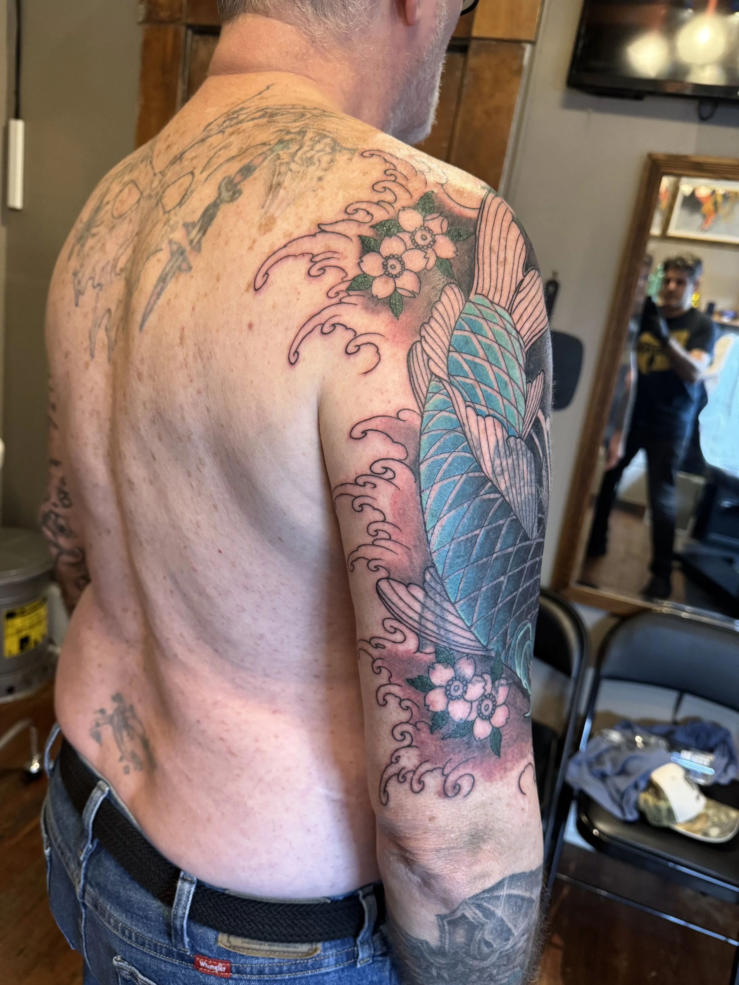 A man with short gray hair and a beard, shirtless, showing his tattooed right shoulder, arm, and chest, with a mirror and a person taking his photo in the background.