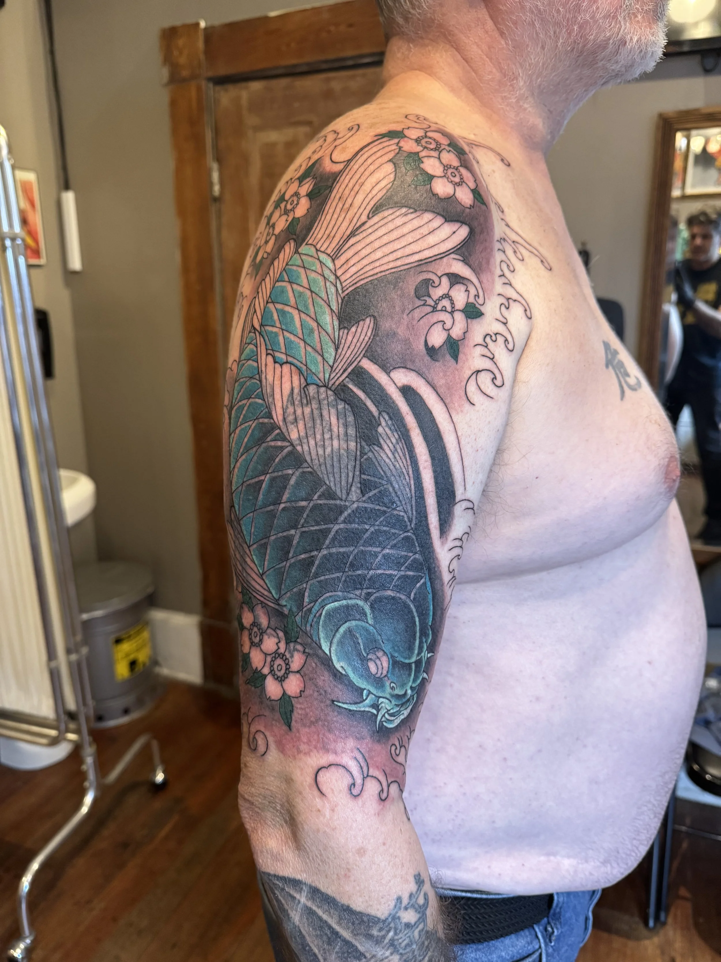 Close-up of a man's upper arm and shoulder tattoo with cherry blossoms, waves, and a fish, in a room with wooden floors and a mirror.