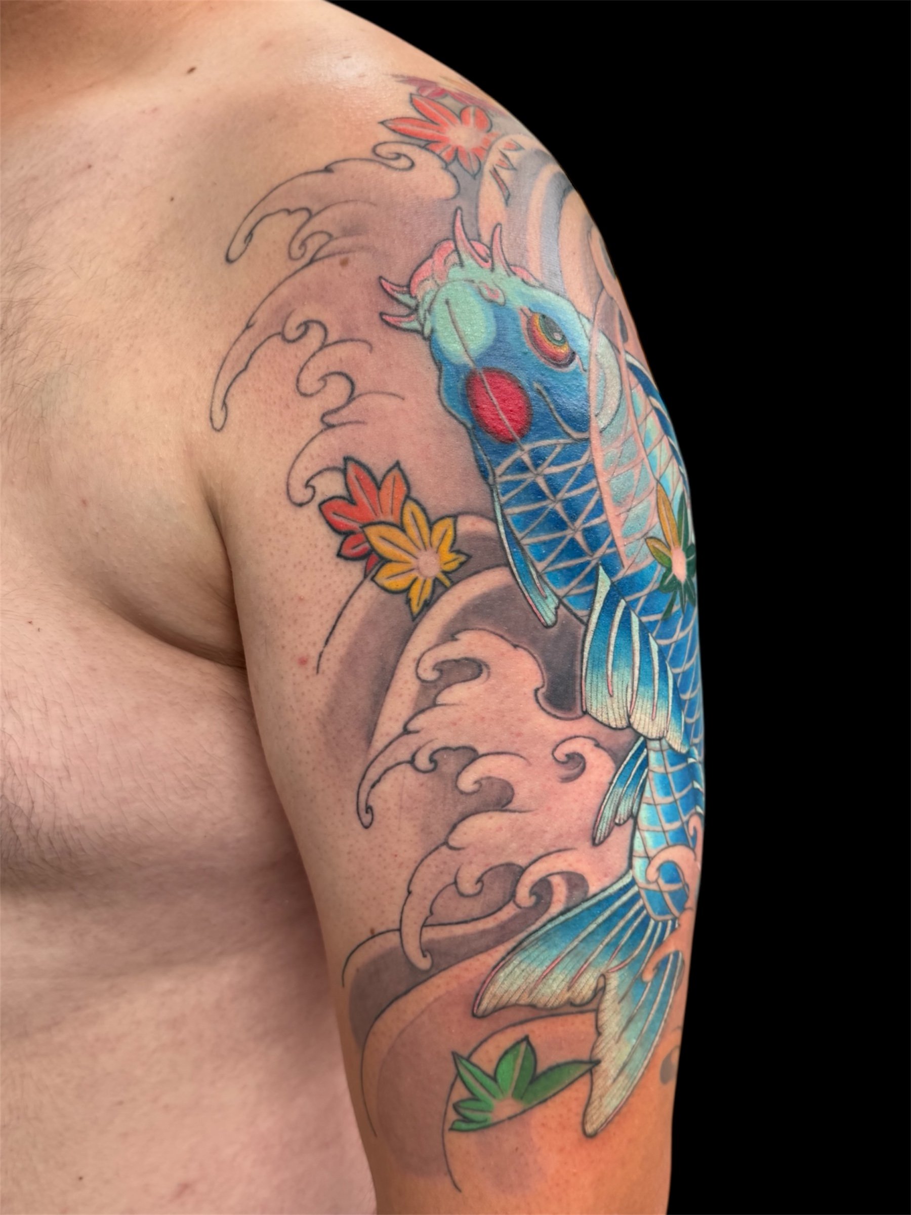 Colorful tattoo of a blue and green koi fish with orange and pink accents, surrounded by stylized waves and pink, orange, and yellow flowers on a person's arm.