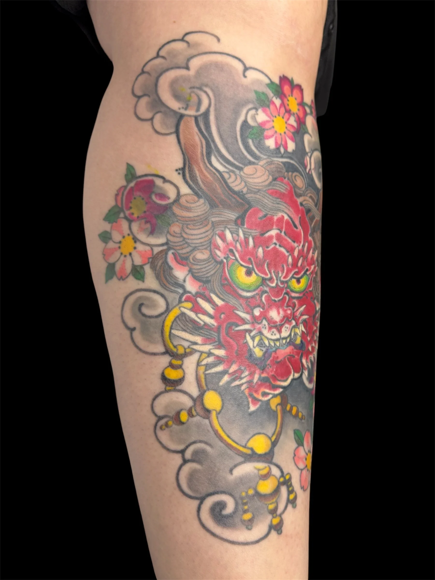 Tattoo of a fierce red demon face with yellow eyes, surrounding flames, and clouds with pink flowers and green leaves on the arm.