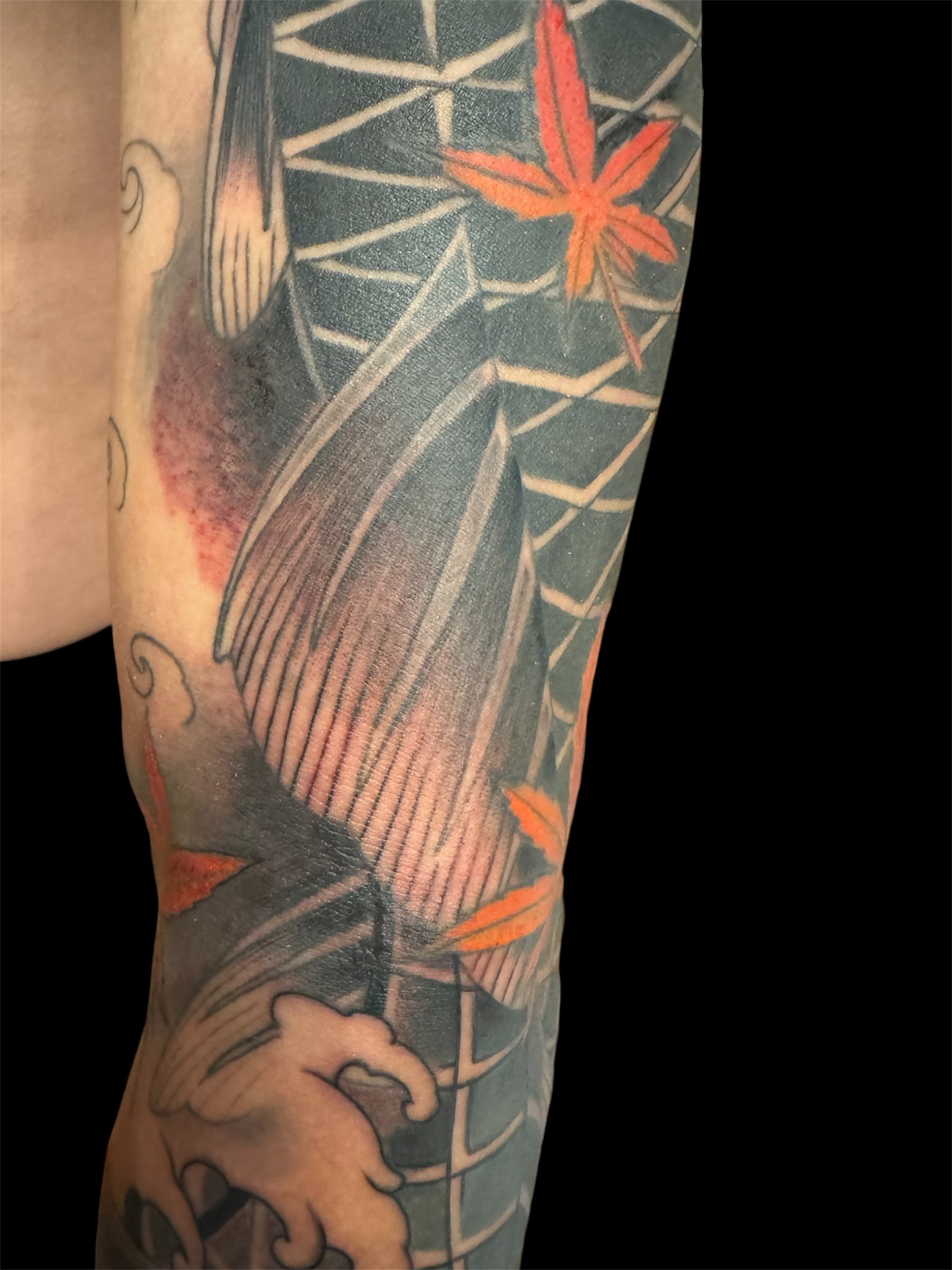 Close-up of a tattoo on an arm, featuring a geometric pattern, a red and orange maple leaf, and cloud designs.