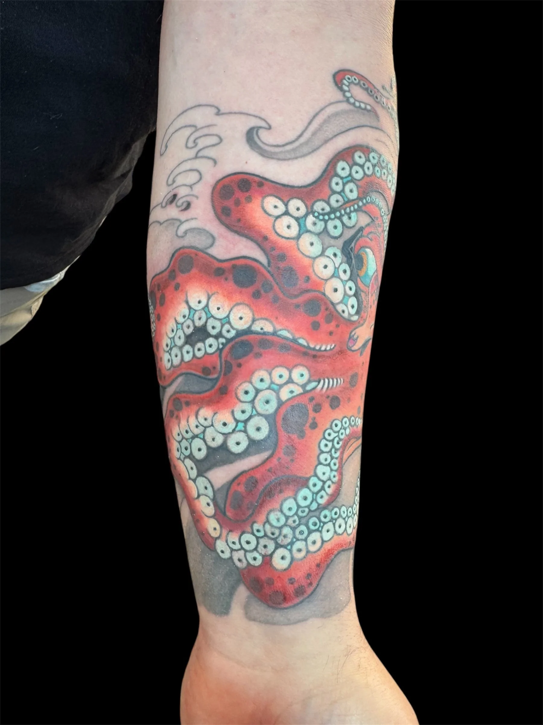 Tattoo of an octopus with red and black coloring, tentacles wrapping around the arm, with detailed suction cups and water splash design, on a person's forearm.