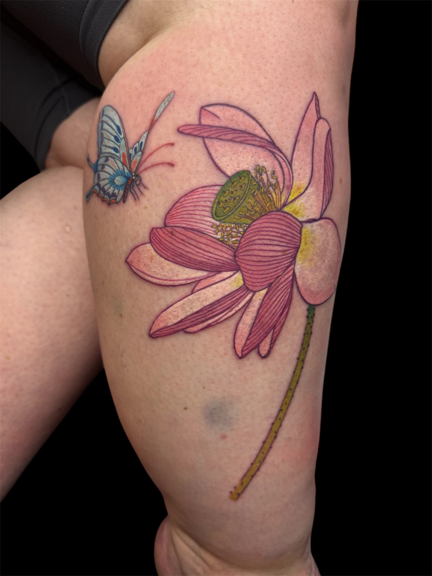 A colorful tattoo of a blue butterfly flying near a pink flower with a yellow stamen on a person's upper arm or shoulder.