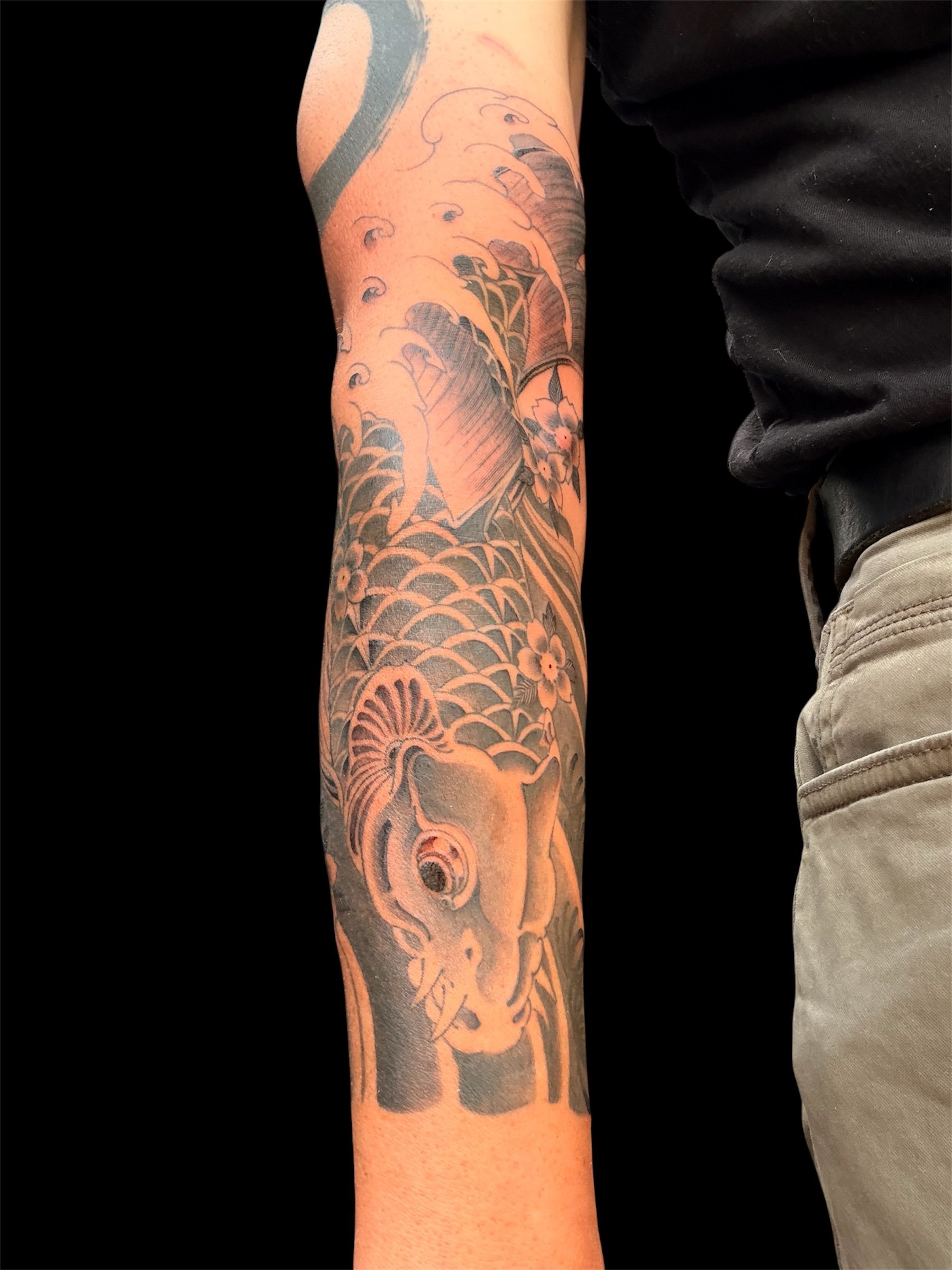 A person's arm with a large black and gray tattoo of a traditional Japanese dragon with floral and wave patterns.