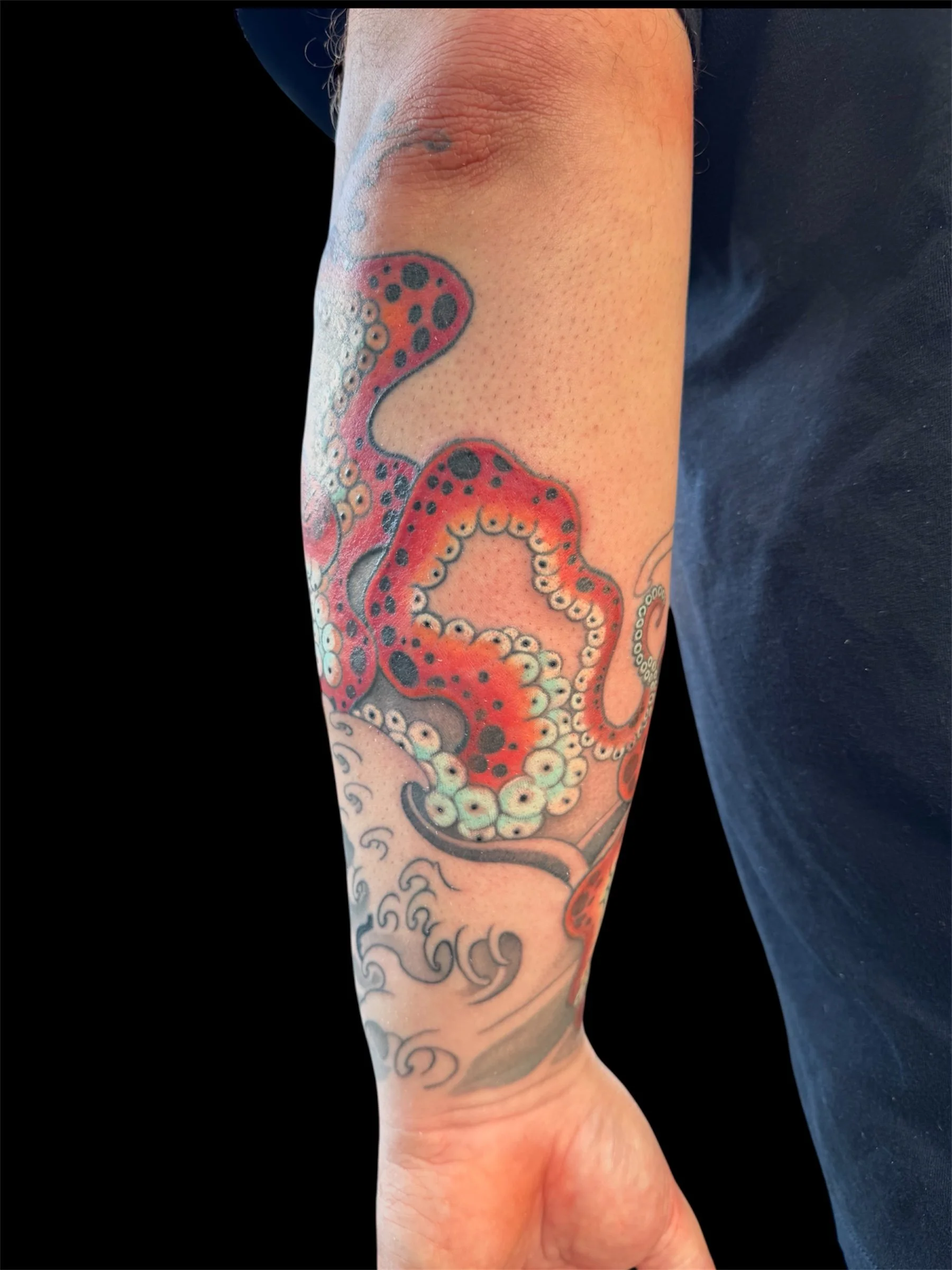 Close-up of a person's arm with colorful tattoo art of an octopus, featuring tentacles and circular patterns, against a black background.