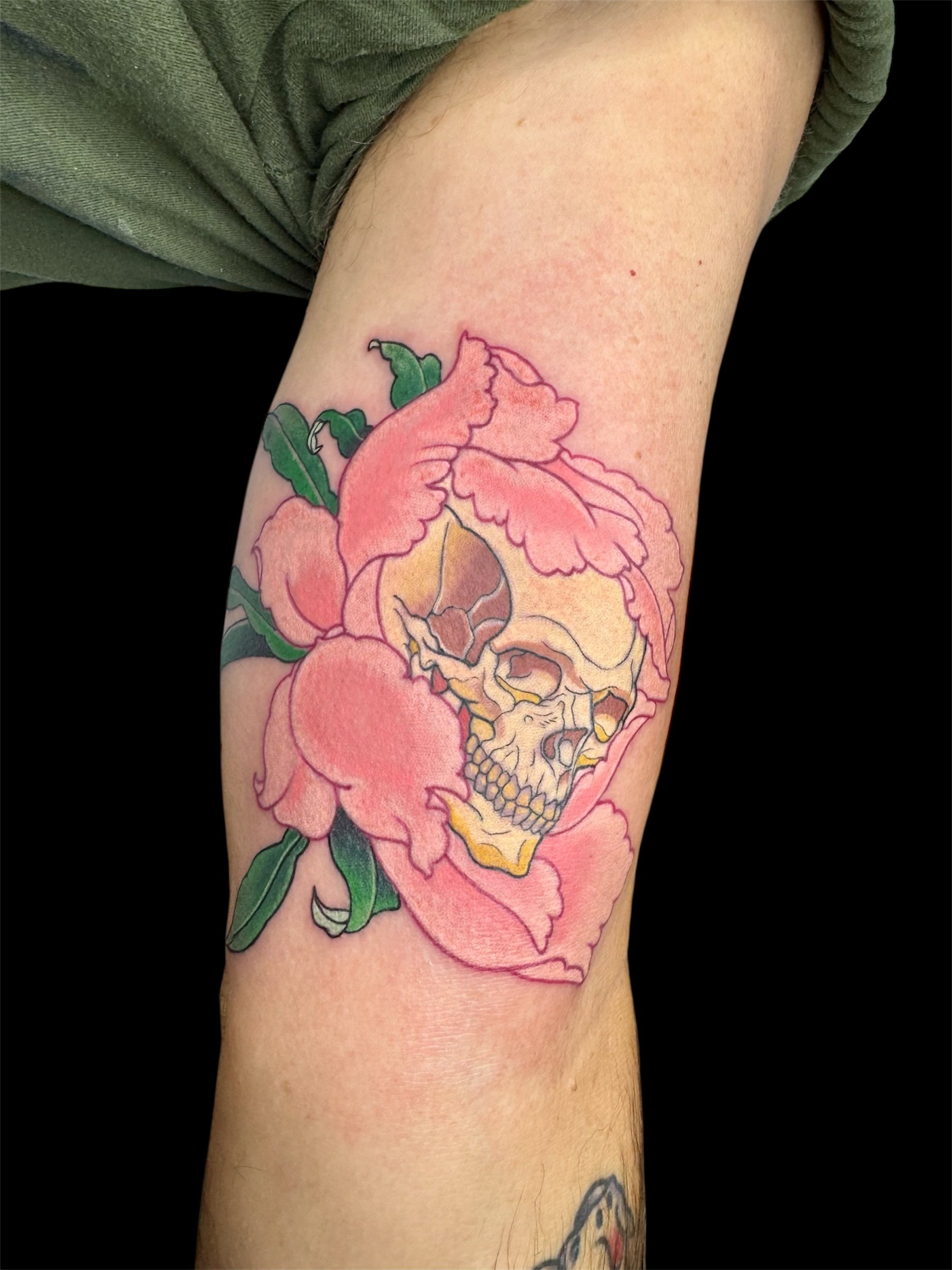 A tattoo of a skull incorporated into a pink flower on a person's upper arm, with green leaves surrounding it.