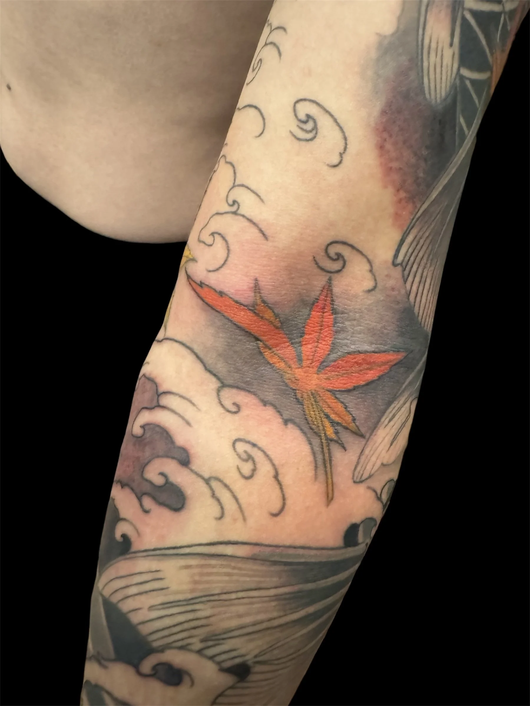 Close-up of a tattooed person's arm featuring a colorful cannabis leaf and swirling cloud patterns.