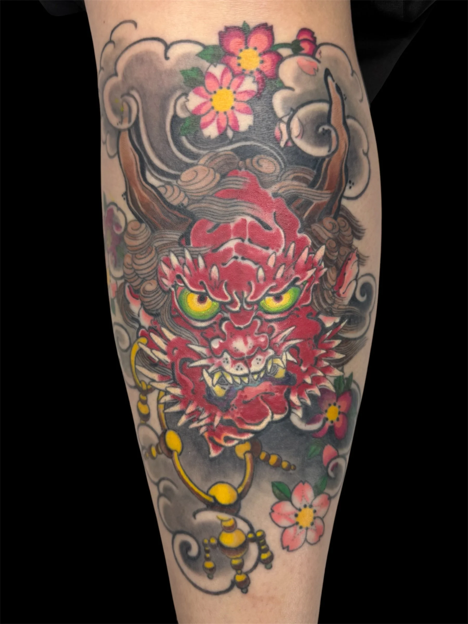 Tattoo of a red and pink floral design with yellow accents, surrounding a stylized face with green eyes and pointed features, all outlined in black, on a person's arm.