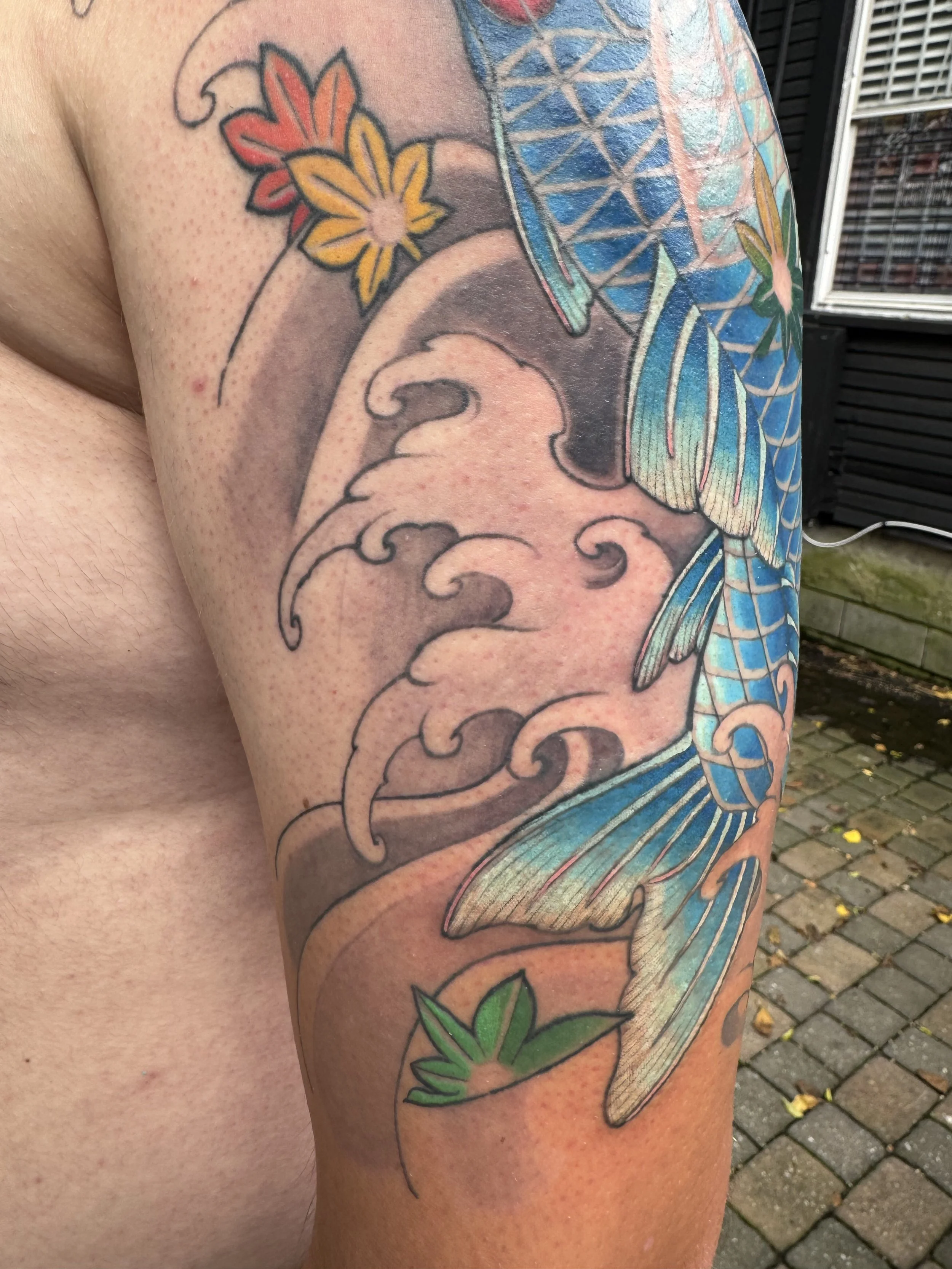 A colorful tattoo featuring a large blue fish surrounded by waves, flowers, and green leaves.