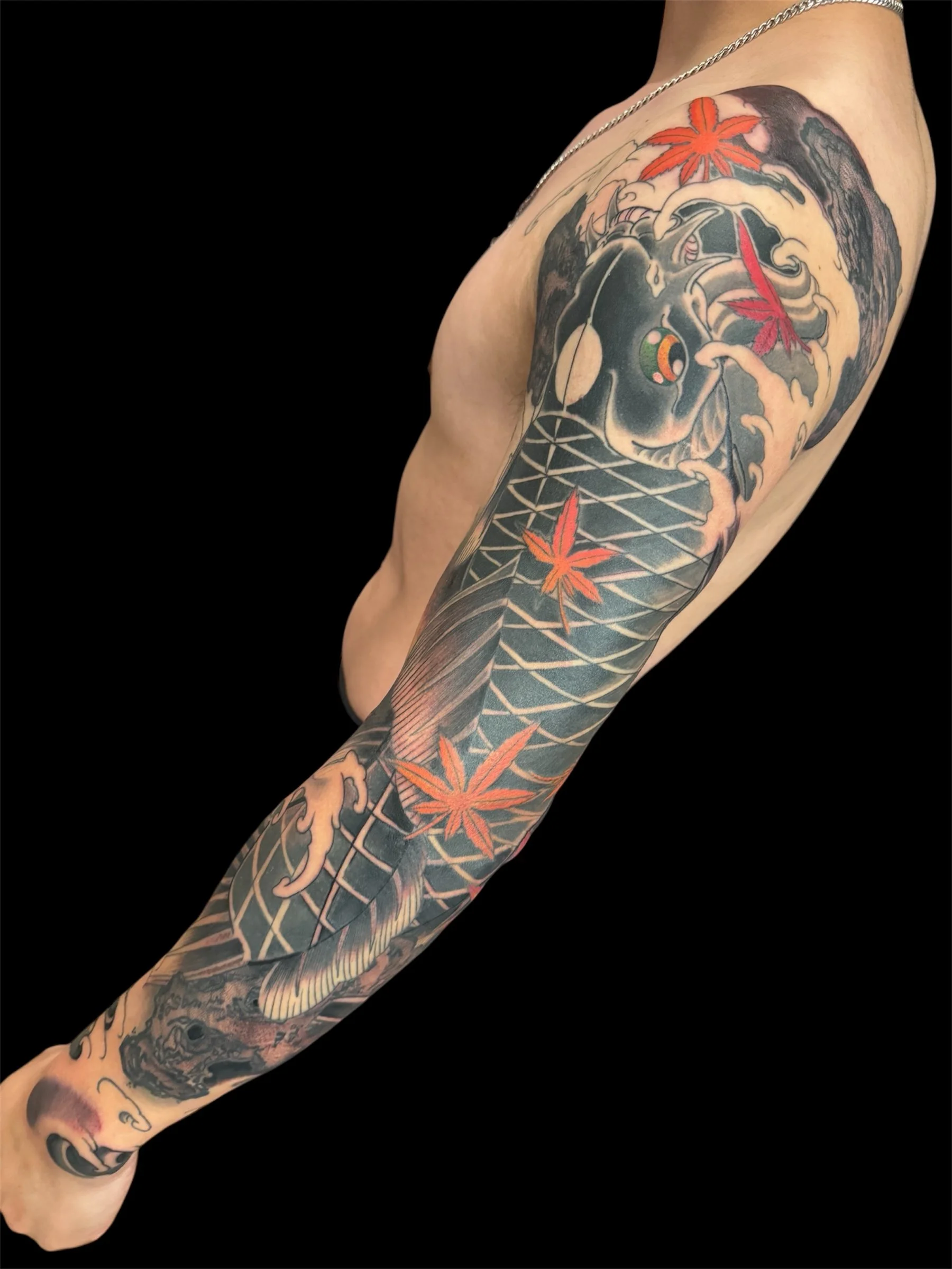 Tattooed arm with a Japanese maple leaf, waves, a tiger, a fish, and geometric patterns against a black background.