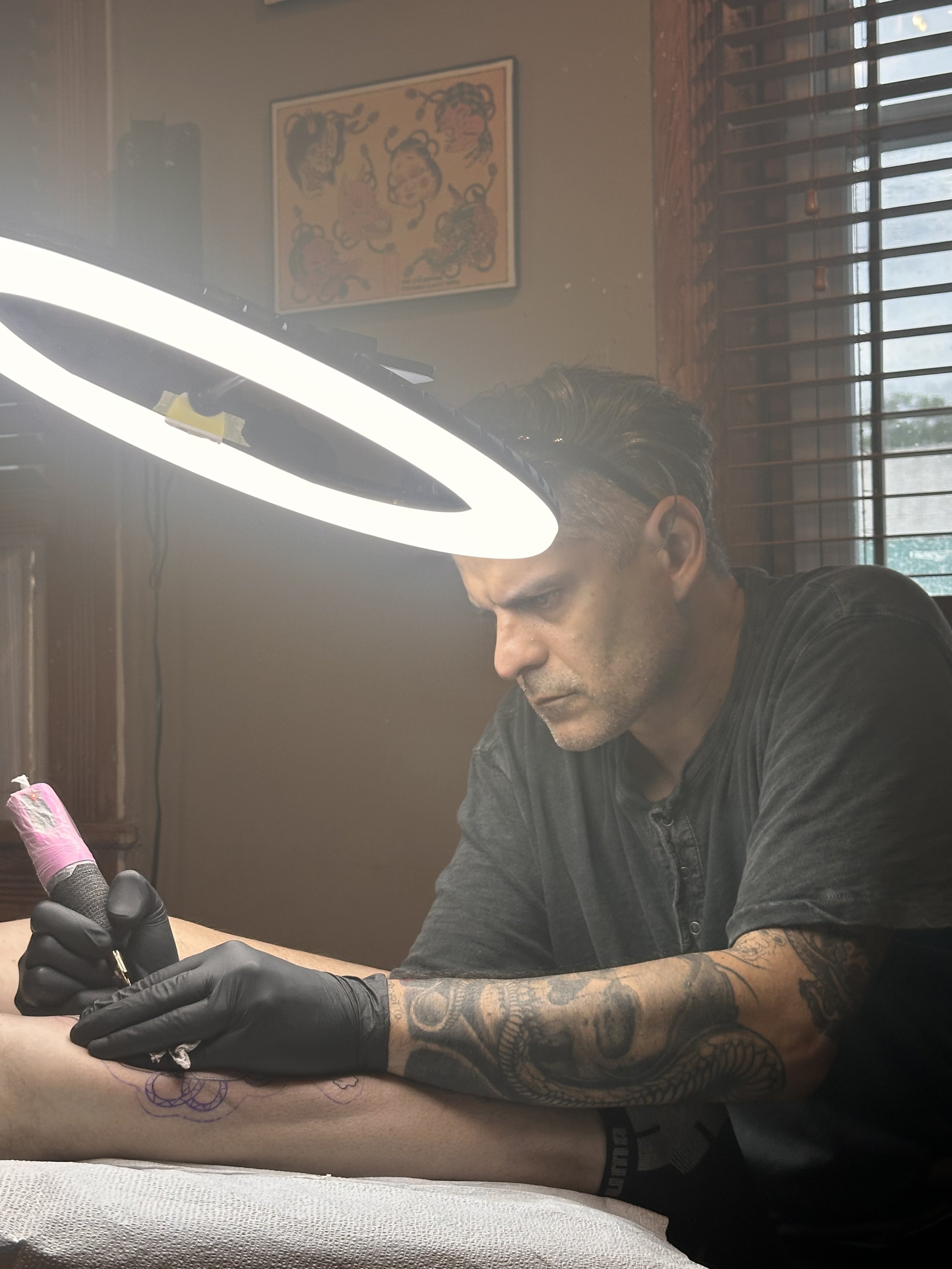 Tattoo artist in black gloves tattooing person's forearm in a studio with wall art and wooden blinds.