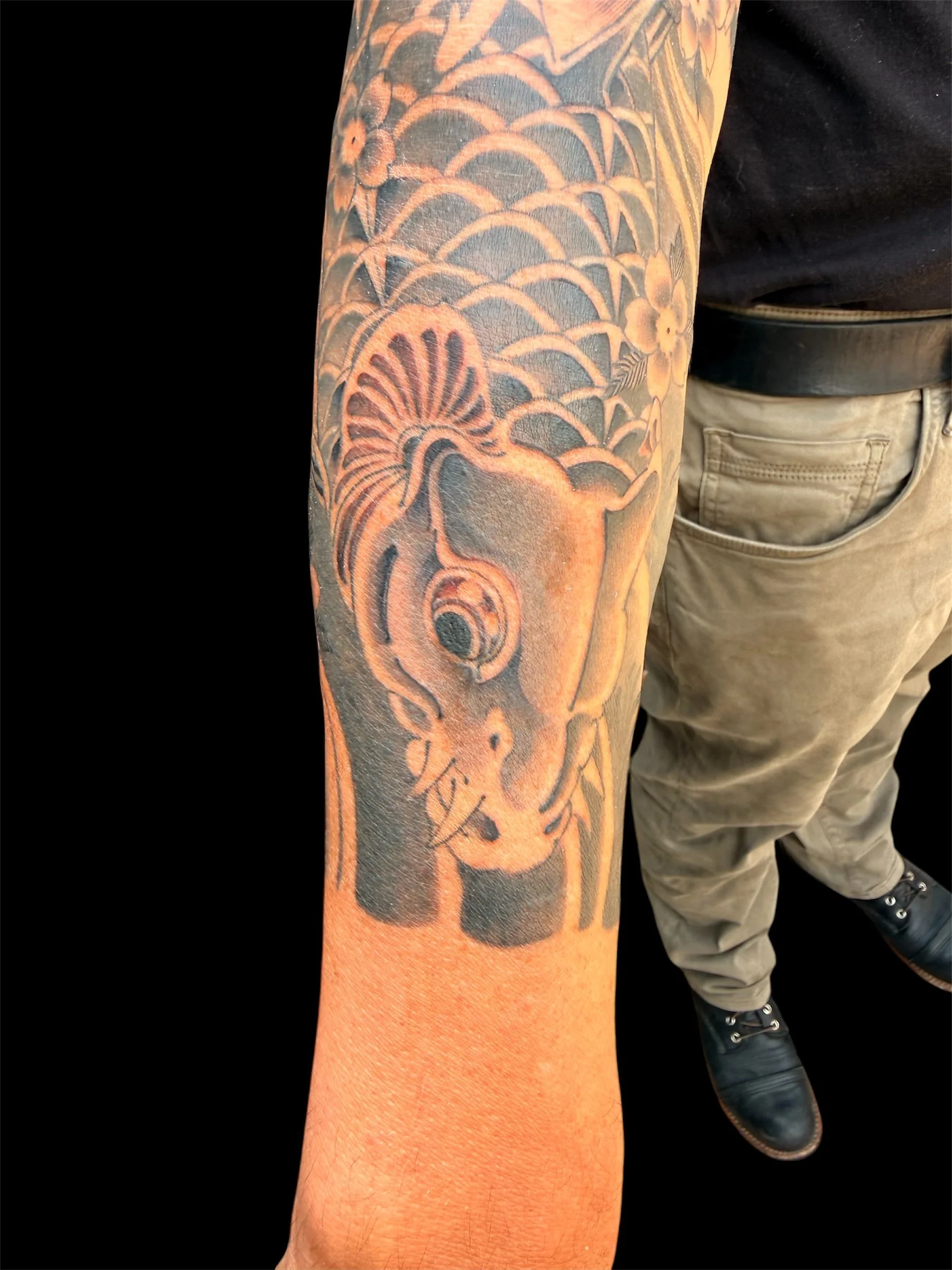 Tattoo on a person's arm features a traditional Japanese-style design with flowers and wave patterns, with a person wearing khaki pants and black boots standing nearby.