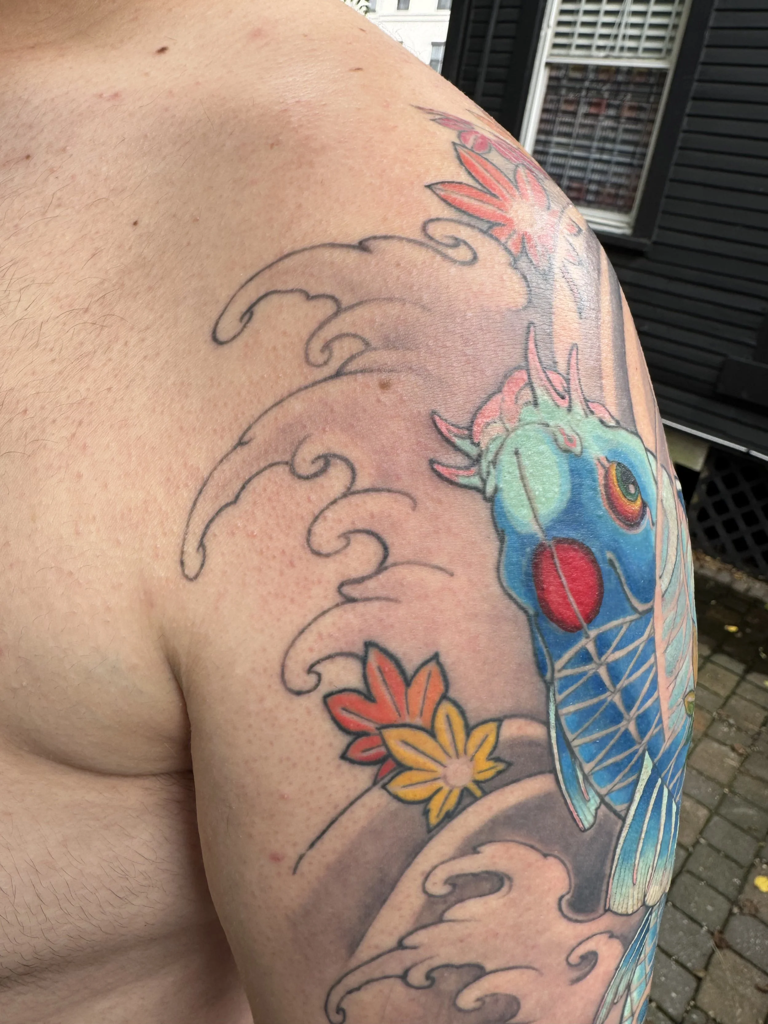 Close-up of a person's shoulder and upper arm with colorful floral tattoos, including red, yellow, and blue flowers and swirling design elements.
