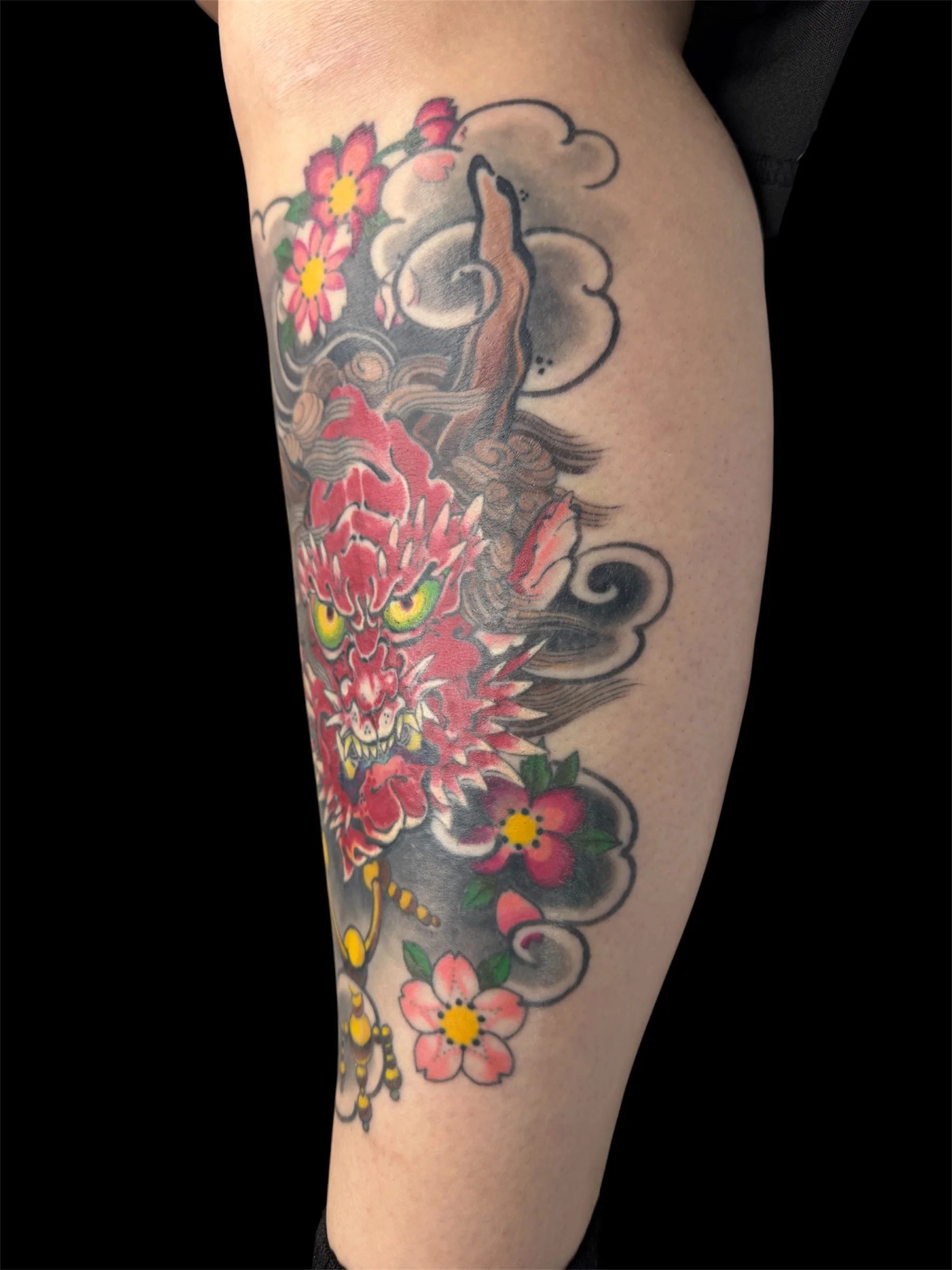 Colorful tattoo of a fierce red and yellow dragon with sharp teeth, surrounded by pink and red flowers with green leaves, on a person's leg.