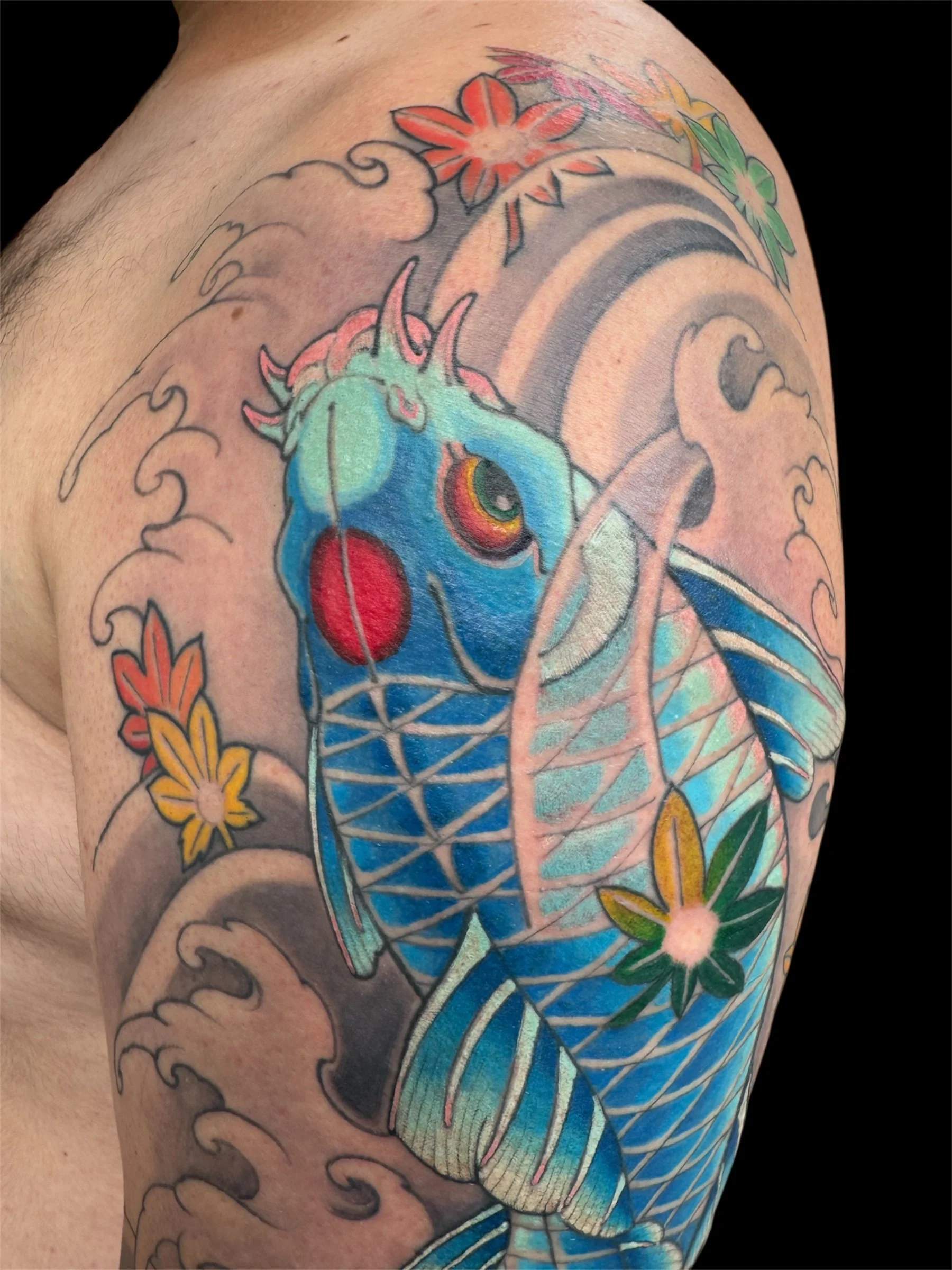Close-up of a colorful tattoo on a person's upper arm, featuring a koi fish with blue and orange scales, surrounded by cherry blossoms and ocean waves.