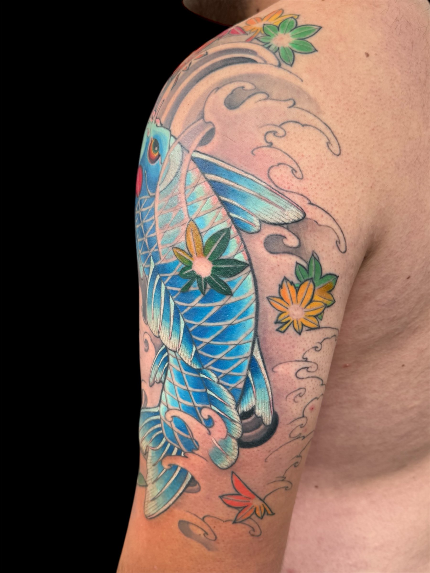 Colorful tattoo of a bird with blue feathers and a red eye on the upper arm, surrounded by floral designs with green, yellow, and orange flowers and decorative wave patterns.
