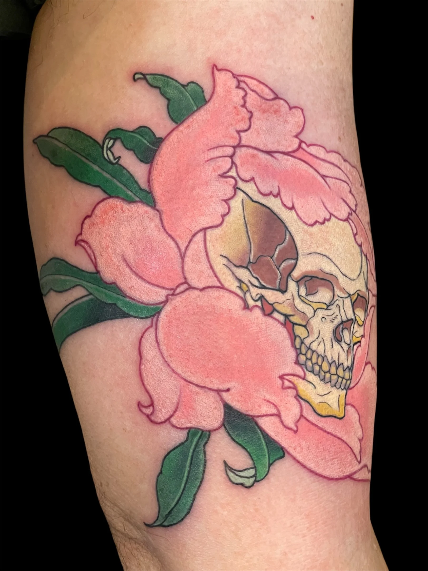 Colorful tattoo of a pink flower with green leaves and a skull incorporated into the flower's center on a person's skin.